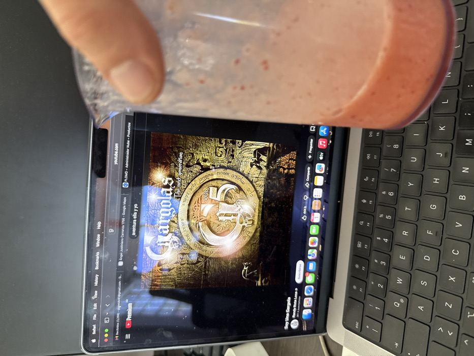 A close-up, eye-level shot captures a human hand gripping a transparent cylindrical container filled approximately one-third with a pinkish-red, textured liquid, consistent with a fruit smoothie, in the foreground. The container and hand partially obscure the right side of an open silver MacBook Pro laptop, which dominates the mid-ground. The laptop screen displays a Safari web browser, prominently showing a YouTube video with the album art for "Gárgolas: The Next Generation - VOL G5" and the channel "Soy Una Gárgola." Multiple browser tabs are open, including one for `youtube.com` and another showing "Angel Justiniano Carranza 1758 - Google Maps," indicating a search for a specific address in the Palermo neighborhood of Buenos Aires, Argentina. The laptop's black QWERTY keyboard is visible below the screen. The background behind the laptop is dark and indistinct, suggesting an indoor environment.