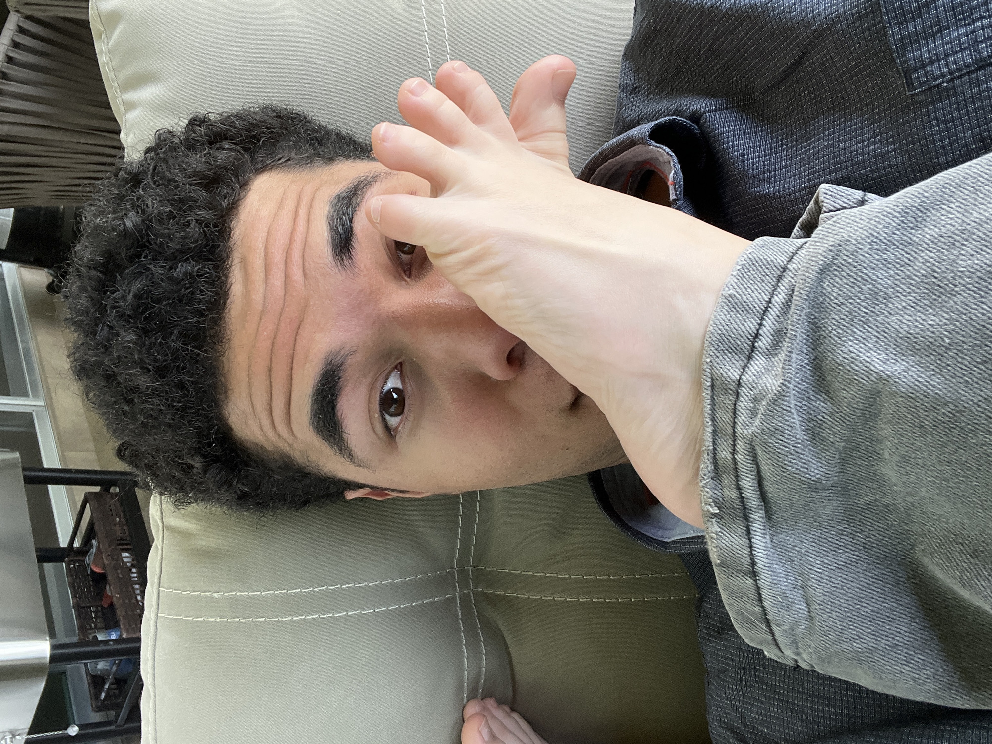 A person with dark curly hair is lying on a beige cushion, with a right foot covering part of the face. The person's face is turned to the side, with one eye visible. The person is wearing a gray denim jacket over a dark gray, patterned shirt. A small portion of a foot is visible at the bottom edge of the cushion. Outside, through a window, a black metal rack is visible. The location is not specified in the image.