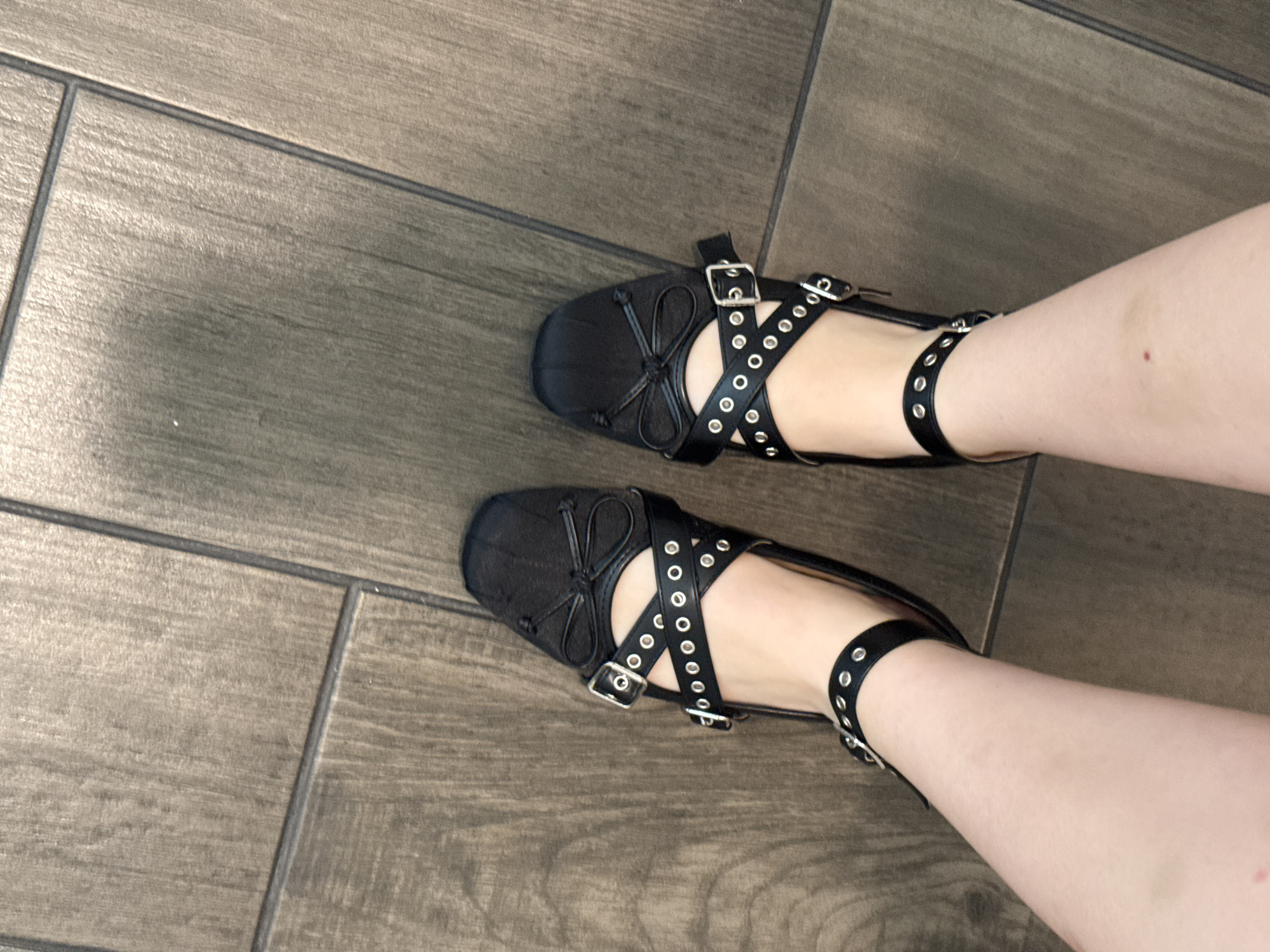 The image shows two feet wearing black ballet-style shoes with multiple straps across the top of the foot and around the ankle, each strap adorned with metallic eyelets and buckled closures. The feet are positioned on a wooden-look tiled floor, with the tiles arranged in a rectangular pattern. The skin tone of the legs and feet is pale, with a small dark spot visible on the right leg. No other people or objects are visible. The location is not identifiable from the image.
