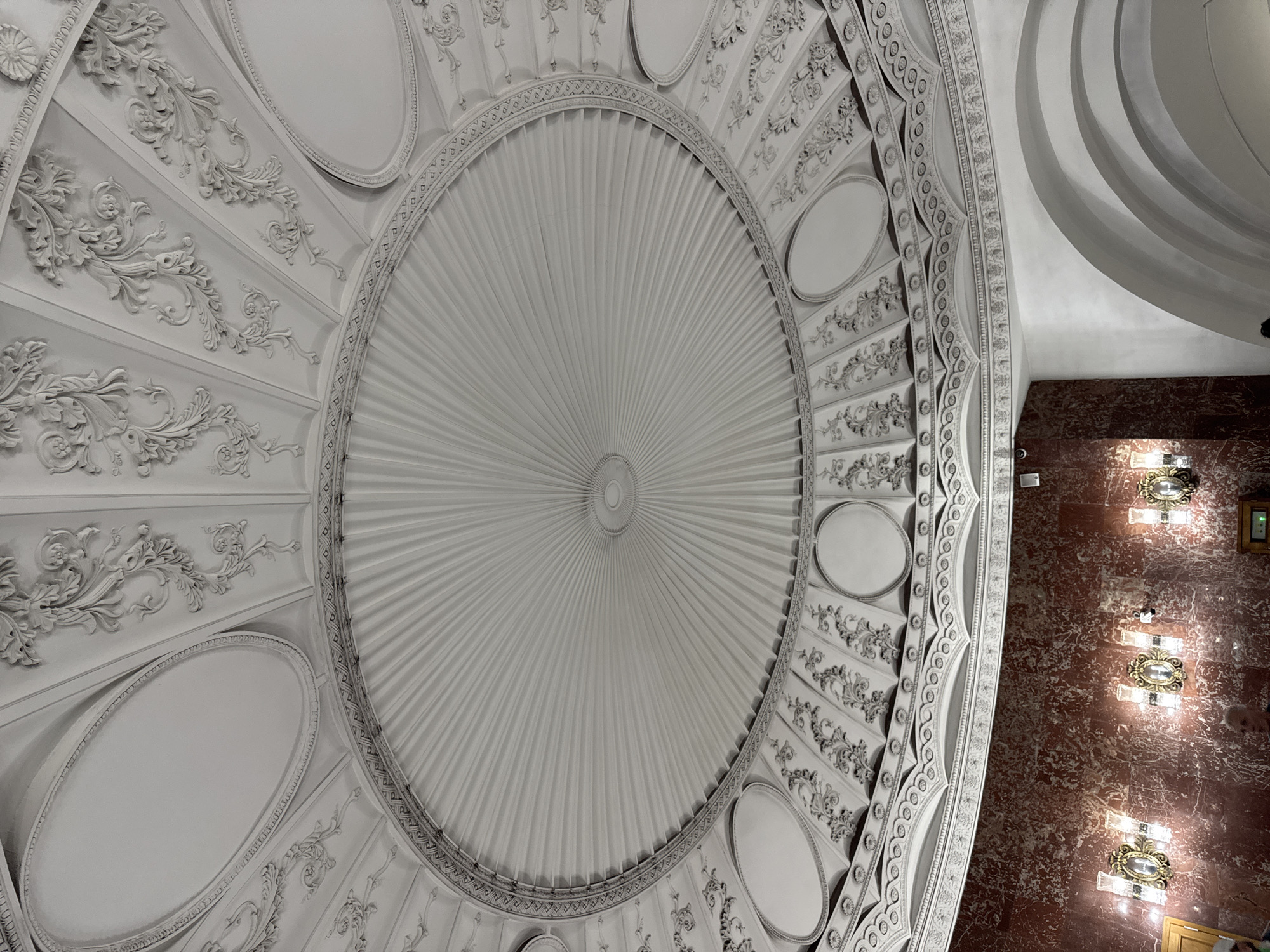 This media file showcases an ornate ceiling with intricate white stucco molding. The ceiling features a large, fluted dome at its center, radiating outwards like rays. Surrounding the central dome are decorative panels with floral motifs and oval recessed areas. A textured, ornate border separates the central dome from the outer decorative elements. To the right, the wall is covered in deep red marble, and several ornate light fixtures with glass and gold accents are mounted on it. The lighting suggests an interior setting, likely illuminated by these fixtures and possibly natural light from unseen sources. The perspective is looking upwards, emphasizing the grandeur and detailed craftsmanship of the architecture. There are no people or visible text in the media file. The style suggests a historical or classic architectural setting, potentially a grand hall, theater, or museum in Moscow, Russia. The ambient lighting and polished marble create an atmosphere of elegance and luxury.