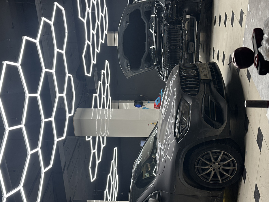 Two cars are in a garage with bright, geometric lights overhead. The first car, a dark gray SUV, is in the foreground, its front end prominent. The second car, partially visible in the background, has its hood open, revealing the engine compartment. Tools and accessories lie nearby on the tiled floor, suggesting the cars are undergoing maintenance or detailing.
