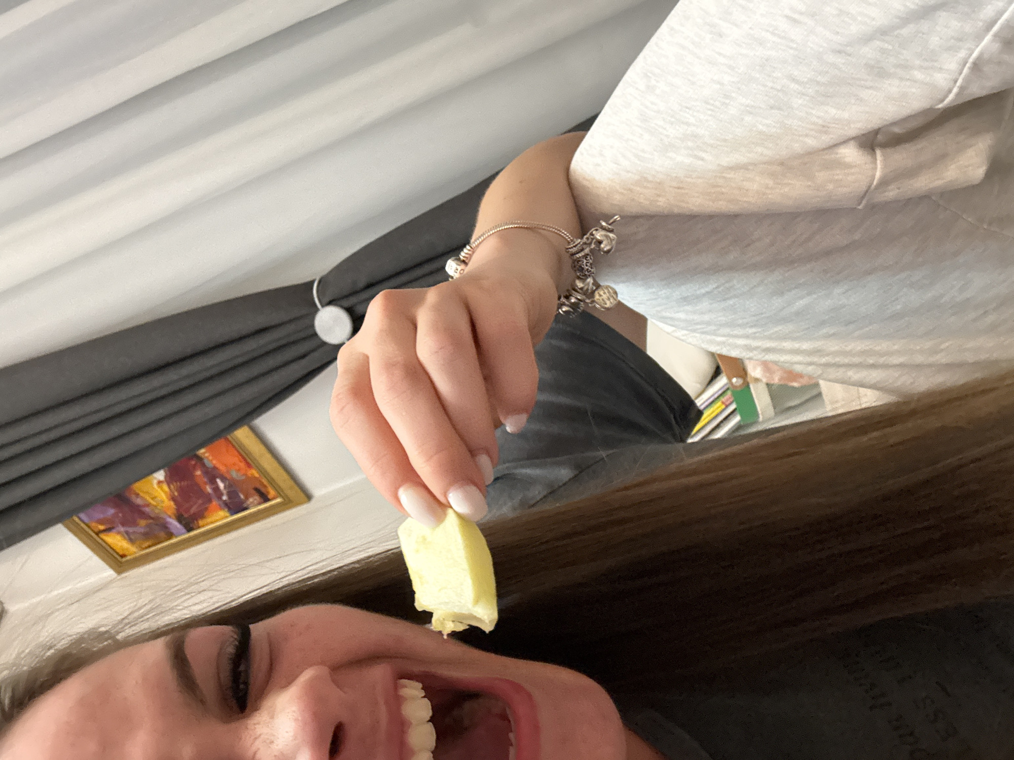 A woman's face is angled toward the camera, with an open mouth and visible teeth. She holds a small, light yellow square of food between her fingers, poised to eat it. The background includes a grey curtain with a white toggle, an unfocused picture frame, and a silver charm bracelet on her wrist. The context does not provide a clear indication of her location in Plovdiv, Bulgaria.
