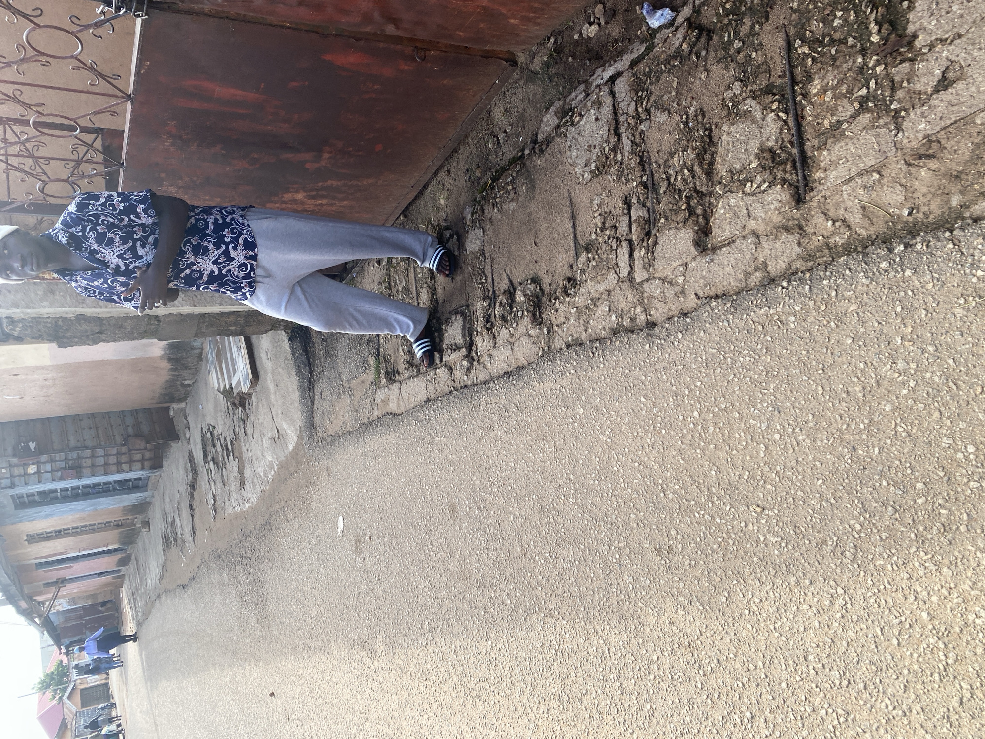 The image depicts a person standing on a cracked concrete walkway next to a road. The person is wearing a dark blue shirt with a white patterned design and light gray sweatpants. They are also wearing dark sandals with white stripes. Behind the person, there is a large, rusty metal surface, possibly a door or gate, and decorative metal bars. The walkway is irregular and shows signs of wear and tear, with some debris and small rocks. The road is paved with asphalt and appears to be a typical street scene. Further in the background, there are buildings and what appears to be a covered walkway or market stall. The lighting suggests it is daytime, with warm, natural light. The scene appears to be set in an urban environment, possibly in Jos, Nigeria, given the context. There is no visible text in the image. The overall impression is a candid street-level shot of an individual in a casual setting.