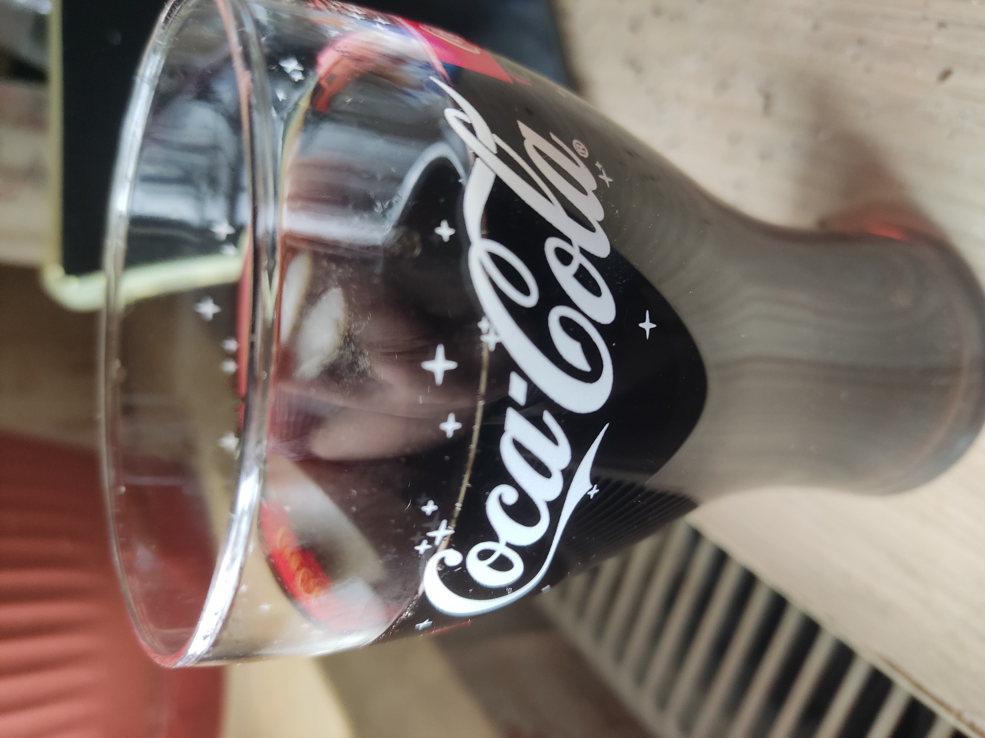 The image shows a Coca-Cola glass. The glass has a black background with the Coca-Cola logo in white. The background has some '+' symbols around the logo. The glass appears to be on a table. The location is Pavlovo, Russia.