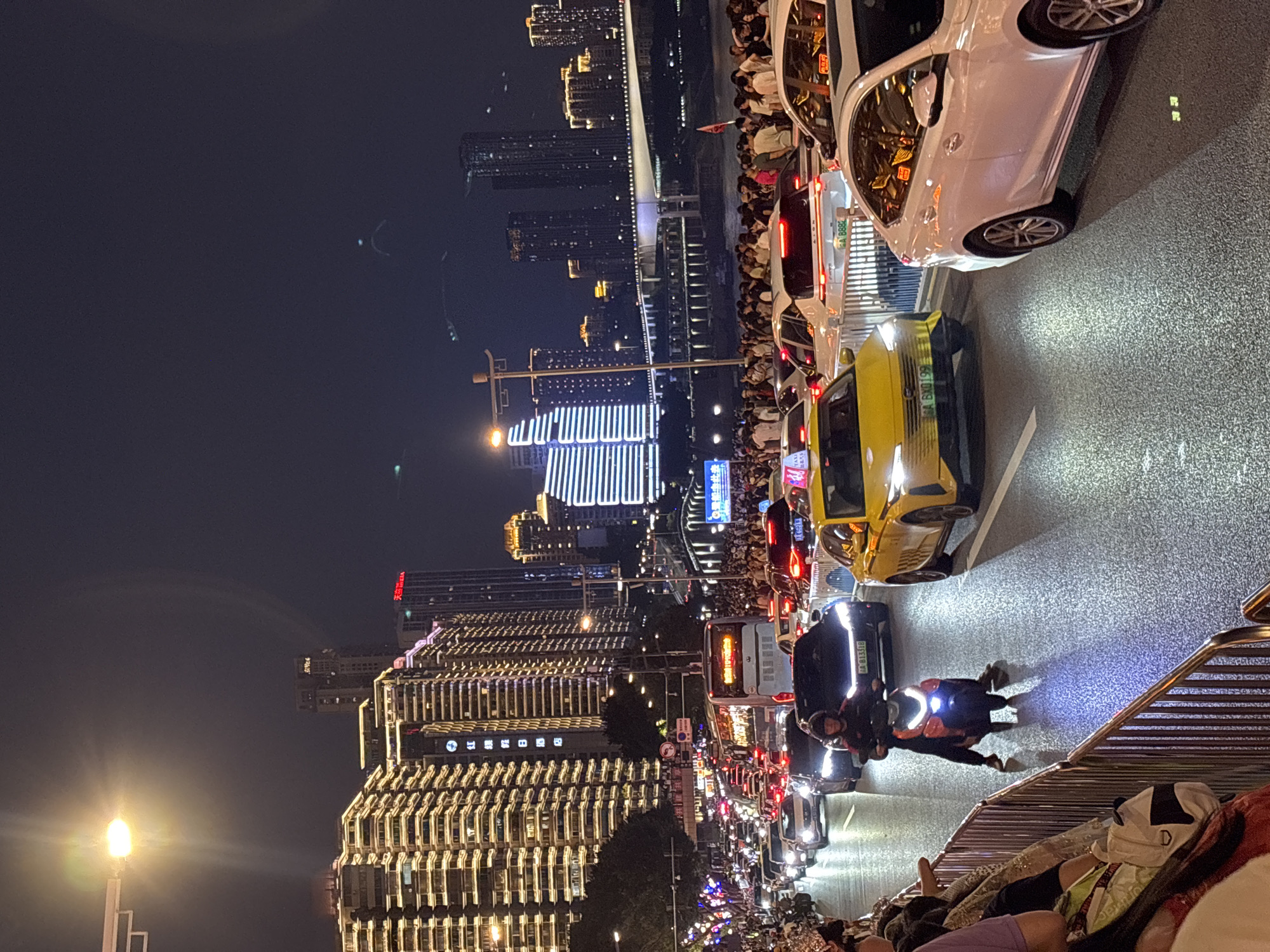 This is a nighttime, eye-level, medium shot of a busy city street in Yuzhong District, China, likely during an event or holiday. The scene is dominated by a traffic jam of cars and a bus on a multi-lane road. Pedestrians are visible on the sidewalks and along a metal barrier in the foreground, many appearing to be spectators. The background features a dense collection of illuminated skyscrapers, some with intricate lighting patterns, and a bridge with lights spanning a body of water. A streetlamp with a bright light is visible in the upper left corner. On the road, a motorcycle with its rider is in motion, and several cars, including yellow taxis and white sedans, are stationary or moving slowly. The overall atmosphere is one of congestion and lively urban nighttime activity.