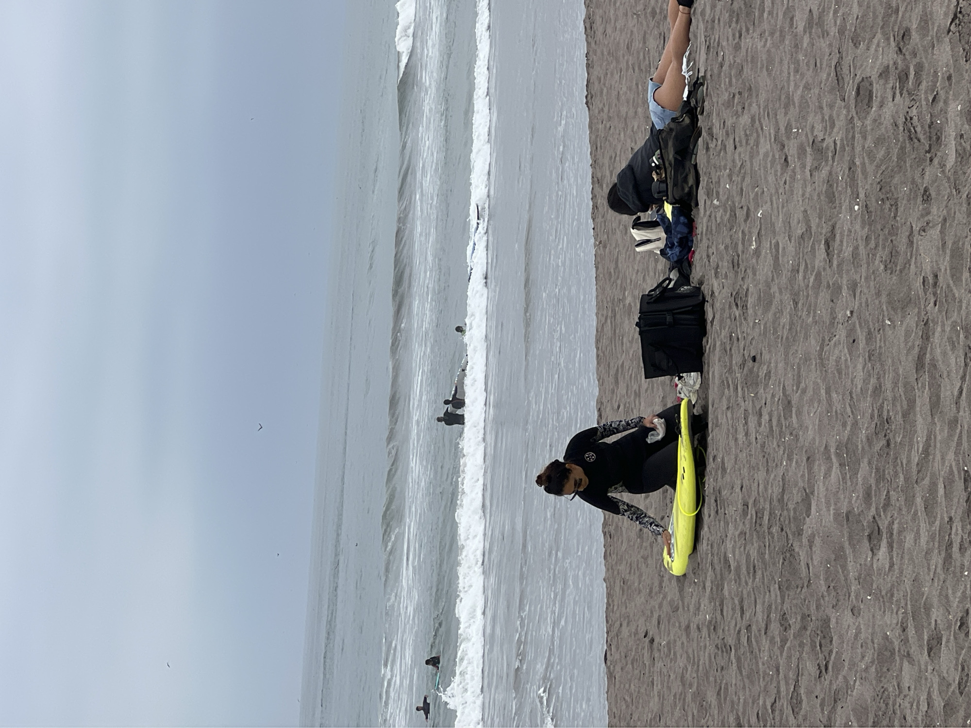 The image shows a beach scene in Arica, Chile. The sky is overcast. The beach has dark sand and the ocean is visible with waves breaking. Several people are surfing in the water. On the beach, a woman wearing a wetsuit is standing and appears to be preparing a yellow bodyboard. Nearby, another person is lying down on the sand with their belongings, including a black bag and clothing.