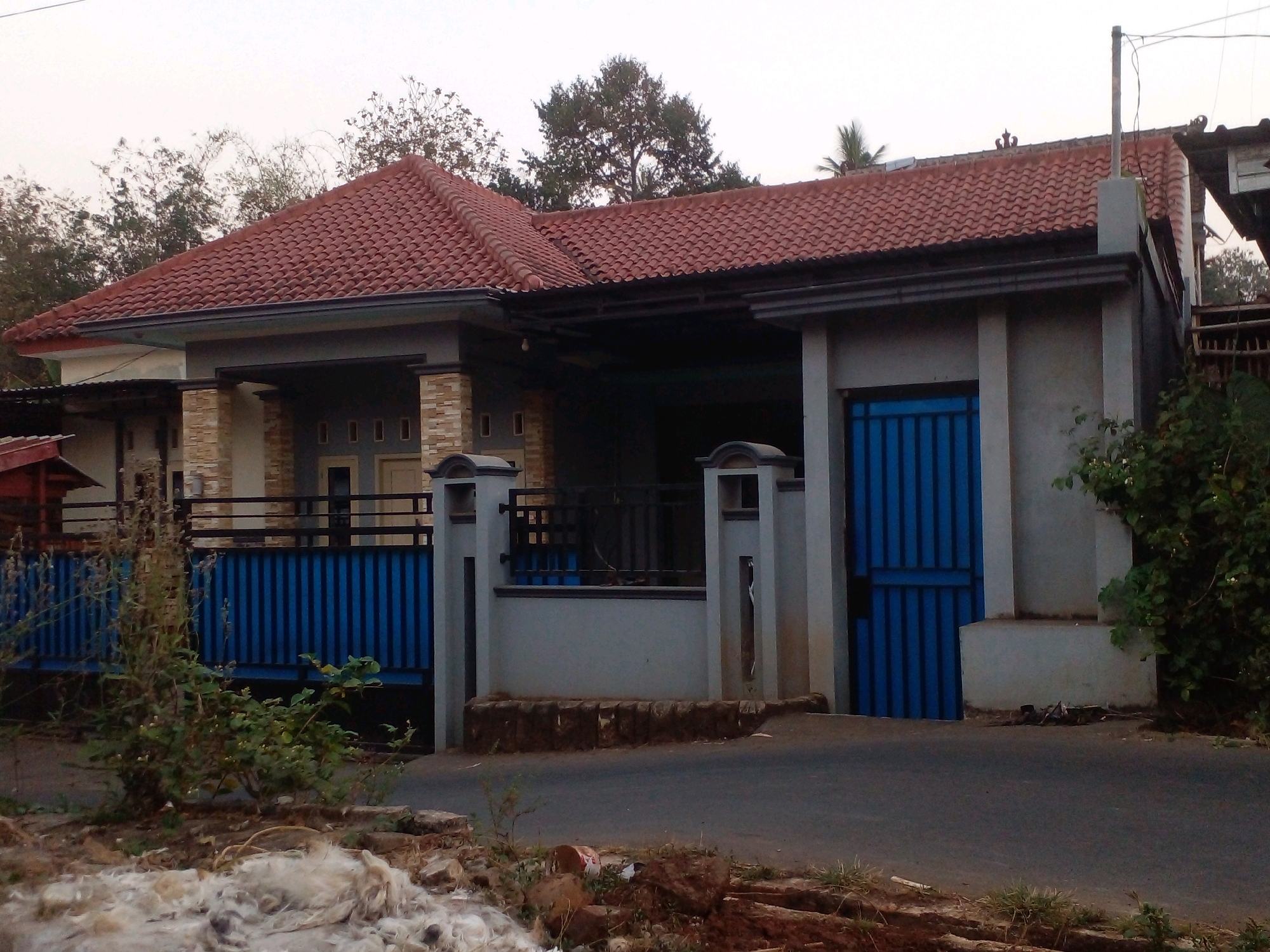 The image shows a single-story house with a red tiled roof, located in Pungangan, Indonesia. The house has a gray facade with stone accents around the entrance. A blue metal gate is visible on the right side of the image, leading to the property. The house is surrounded by some greenery and a paved road. The sky is overcast. There are no people or animals visible in the image. The scene appears to be a residential area.