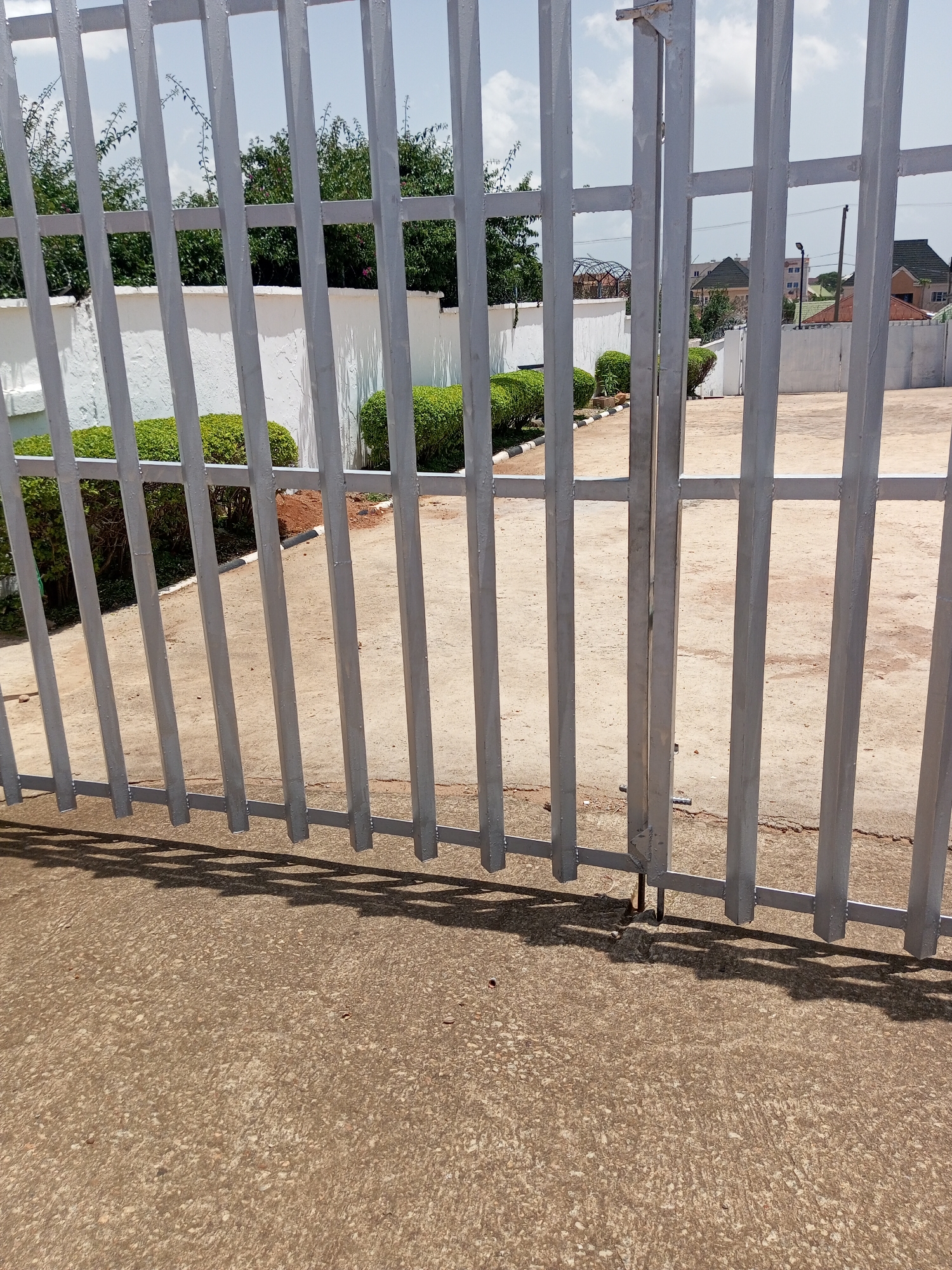 A metal gate in Bukuru, Nigeria, obstructs the view of a paved area behind it, lined with white walls and manicured bushes. The gate is constructed of vertical metal bars, painted grey, connected by horizontal beams, and has a slight shadow cast upon the concrete ground in front of it. In the background, beyond the white walls, are glimpses of trees, buildings with green roofs, and a clear sky with some clouds. The paved area appears to be part of an outdoor space, possibly a courtyard or a yard.