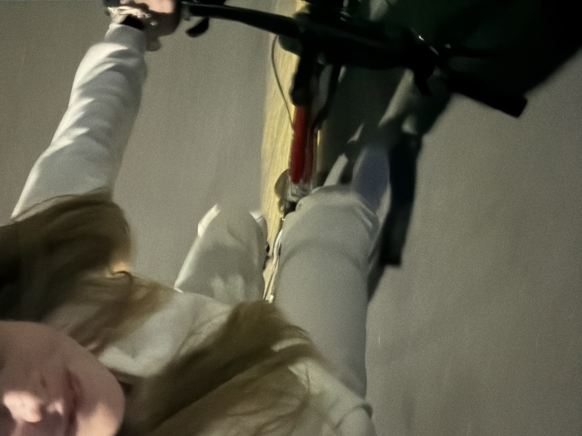 An overhead, slightly blurry, and grainy image captures a person riding a bicycle at night in Izhevsk, Russia. The perspective is from above the rider, looking down towards their torso, legs, and the front section of the bike.
The primary subject is the individual, who appears to be wearing a white long-sleeved top and light-colored pants. Long, light brown or blonde hair is visible, falling over their chest. One hand, adorned with what looks like a wristband or watch, is visible gripping the dark handlebars of the bicycle.
The bicycle itself features dark handlebars and a distinct reddish section of its frame visible towards the front. The scene is set in low-light conditions, strongly suggesting it is nighttime. Illumination is dim, possibly from artificial sources, casting muted tones across the image. A faint, elongated streak of yellowish light is visible on the ground next to the bicycle, which could be a reflection or an illuminated part of the path.
The image's notable details include its distinct grainy texture and slight motion blur, characteristic of photography taken in challenging low-light conditions and possibly while in motion. The overall atmosphere is quiet and dark. No specific weather conditions can be discerned, nor are any emotions clearly visible from the partial view of the person. There is no visible text in the image.