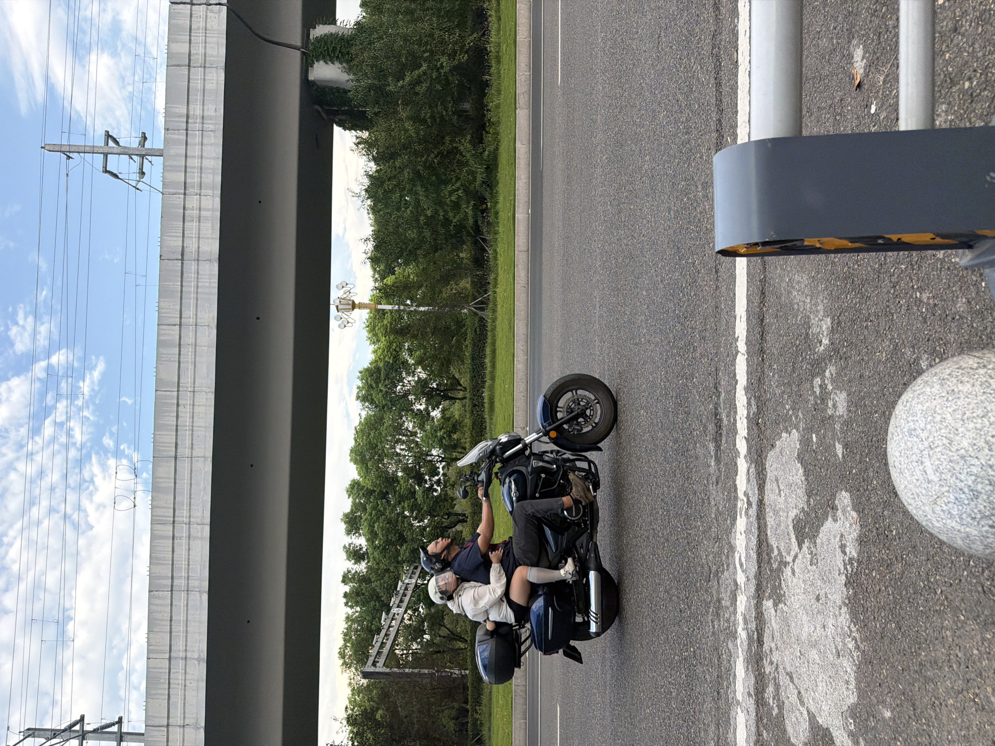 In the city of Jiangjin, China, a man and a woman are riding a motorcycle on a road under an elevated highway. The man is driving, wearing a dark shirt, dark pants, and a helmet. The woman is a passenger behind him, wearing a light-colored jacket and shorts, with a light-colored helmet. They are both wearing helmets. The motorcycle is dark blue and has saddlebags. The background features lush green trees, a streetlight, and the underside of the highway infrastructure with visible electrical lines. The road surface is asphalt with white lane markings. A grey concrete barrier and a decorative stone bollard are visible in the foreground. The sky is partly cloudy.