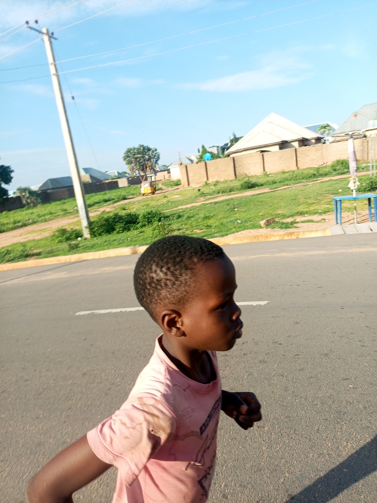 A young child, appearing to be of African descent and with dark skin and short, curly hair, walks on an asphalt road. The child is wearing a light pink t-shirt that appears slightly stained. The child's right arm is bent at the elbow, and their left arm is positioned forward, suggesting movement. The child's head is turned to the right, and their gaze is directed away from the camera.
The background consists of a grassy area with scattered bushes and trees. Several buildings, likely homes with light-colored roofs and brick or concrete walls, are visible beyond a low, tan-colored fence. A utility pole with wires stretches upwards on the left side of the frame. In the distance, a yellow three-wheeled vehicle is parked near a tree. A blue table with a white umbrella or shade is situated on the far right. The sky above is clear and blue, with a few wispy white clouds. The scene is likely an outdoor environment in Tempure, Nigeria.
