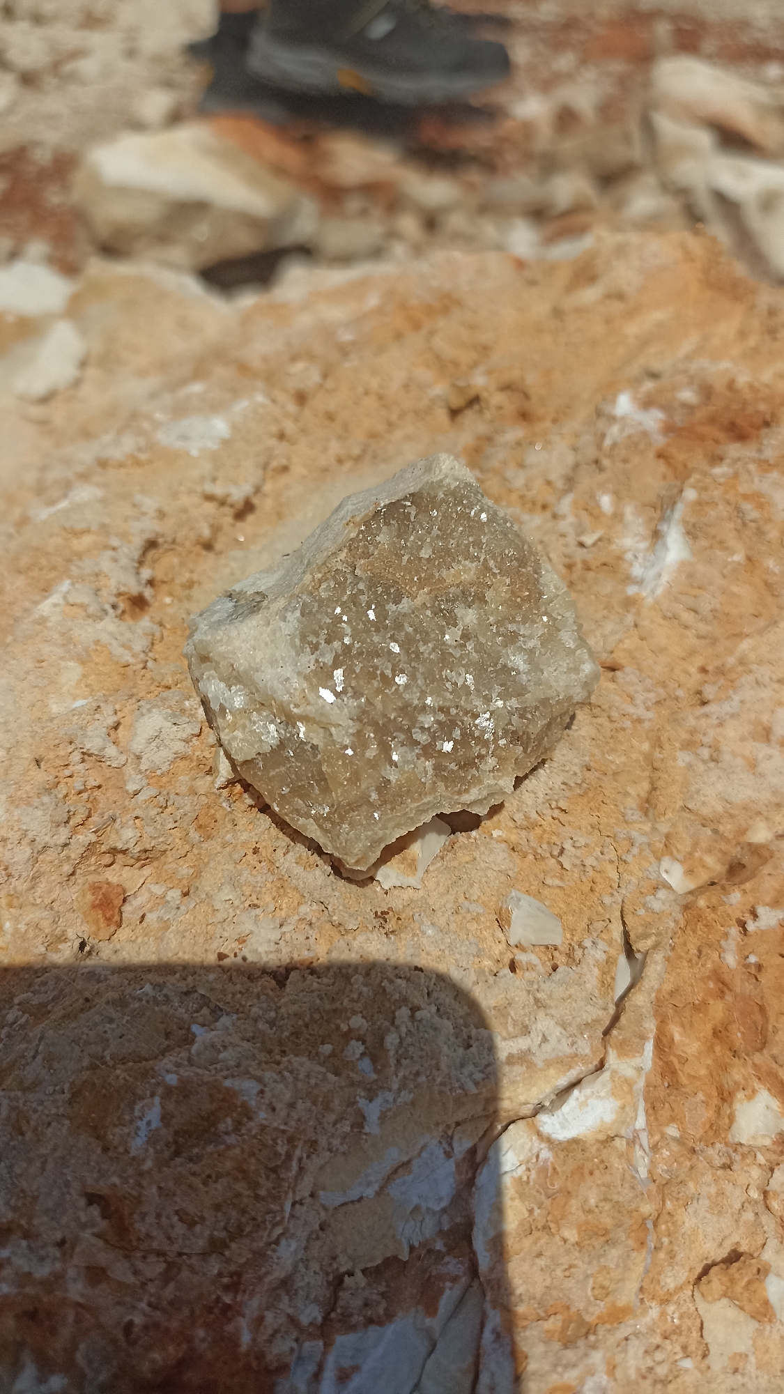 The image shows a beige, cube-shaped mineral specimen with visible inclusions resting on a tan, rocky surface. The mineral exhibits a rough texture and appears to contain reflective flakes, possibly mica. The surrounding rock is fractured and layered, with variations in color ranging from light tan to reddish-brown. The environment appears to be a natural rock formation or outcrop. A blurry shoe is visible in the background. The location is Unknown, Unknown.