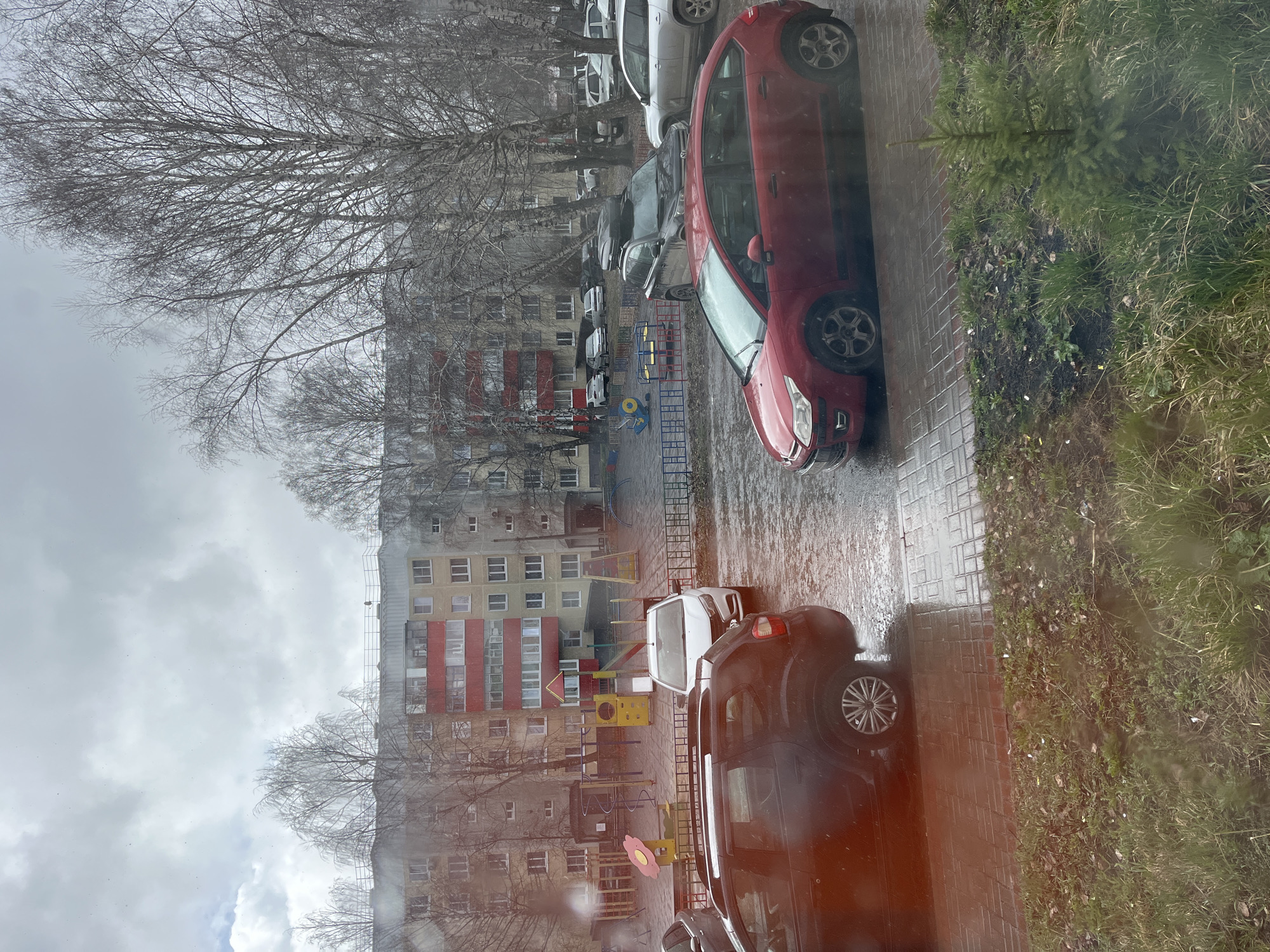 The image depicts an overcast scene in Usman, Russia. Visible from an elevated position, possibly a window, the view includes a residential area. A multi-story apartment building with a red and yellow facade faces a playground. The playground features typical play equipment, including slides and climbing structures. Parked cars line the street, including a red hatchback and several other vehicles. Bare trees are visible, suggesting a late autumn or winter season. The ground appears wet, possibly from recent rain or snow. The image is slightly blurry, perhaps due to condensation on the window. No people are directly visible in the frame.
