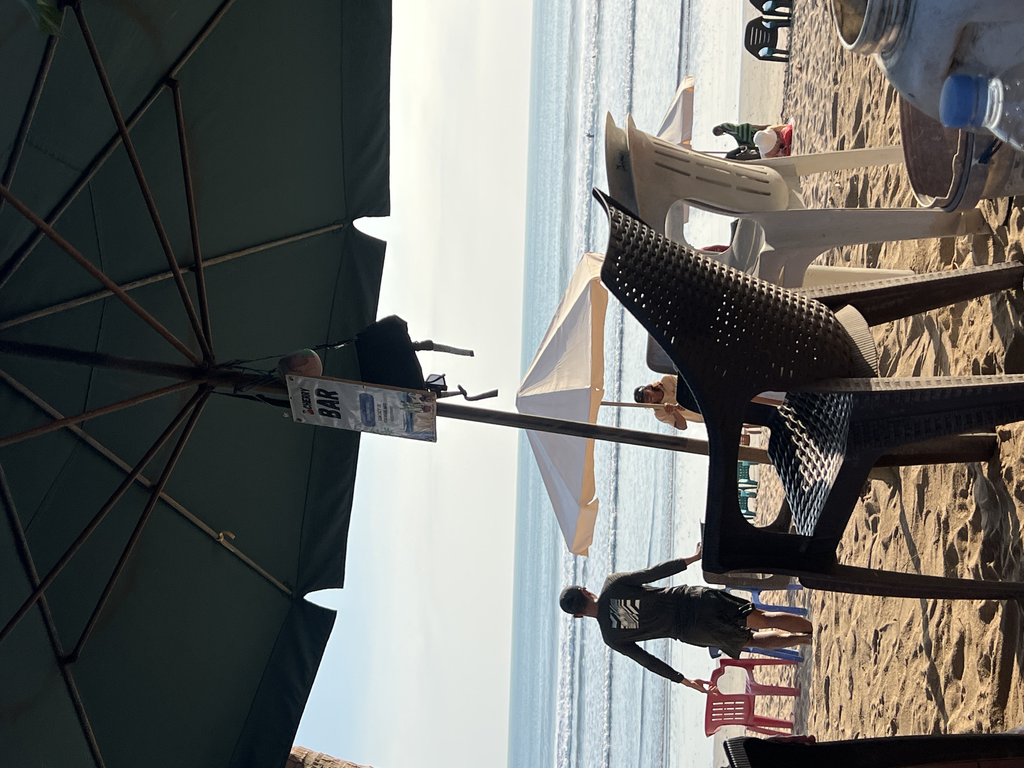The image depicts a beach scene in Kuta, Indonesia, with a large dark green umbrella providing shade. A sign hanging from the umbrella reads "CHERRY BAR". In the foreground, there are several plastic chairs, one of which is a dark brown woven-style chair in the center. Beyond the chairs, a person wearing a dark long-sleeved shirt and shorts stands on the sand, facing the ocean. Several colorful plastic chairs are visible behind this person, including red and blue ones. The ocean in the background shows small waves, and there are a few people in the water. To the right of the central figure, a white umbrella is set up, and another person is seated under it. The beach is sandy, with footprints and patterns from the sand. The overall atmosphere suggests a relaxed beachside establishment.