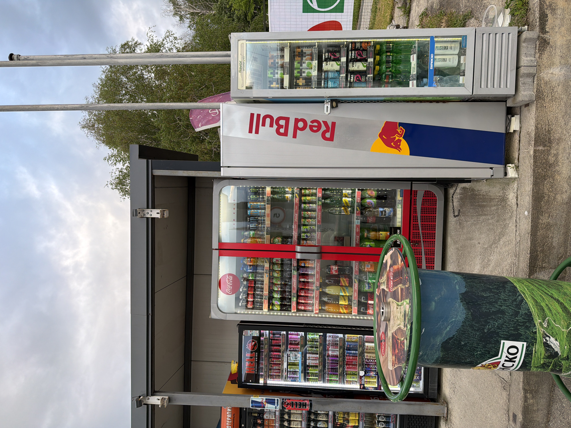 This outdoor shot features an array of vending machines and beverage coolers adjacent to a covered structure. A cooler bearing the "Red Bull" logo is prominently visible, with a Coca-Cola-branded machine directly beneath it. Other refrigerators house various beverages and energy drinks. To the right, a cylindrical object wrapped in a green and blue graphic obscures part of the vending machines. A sign with Cyrillic text is visible near the base of the machines, along with a "HELL" energy drink advertisement. Based on the signage ("ПИРИНСКО," "PELISTERKA," "CKO") and language (Cyrillic text), this scene is likely located in Bulgaria, specifically, Panagyurishte.