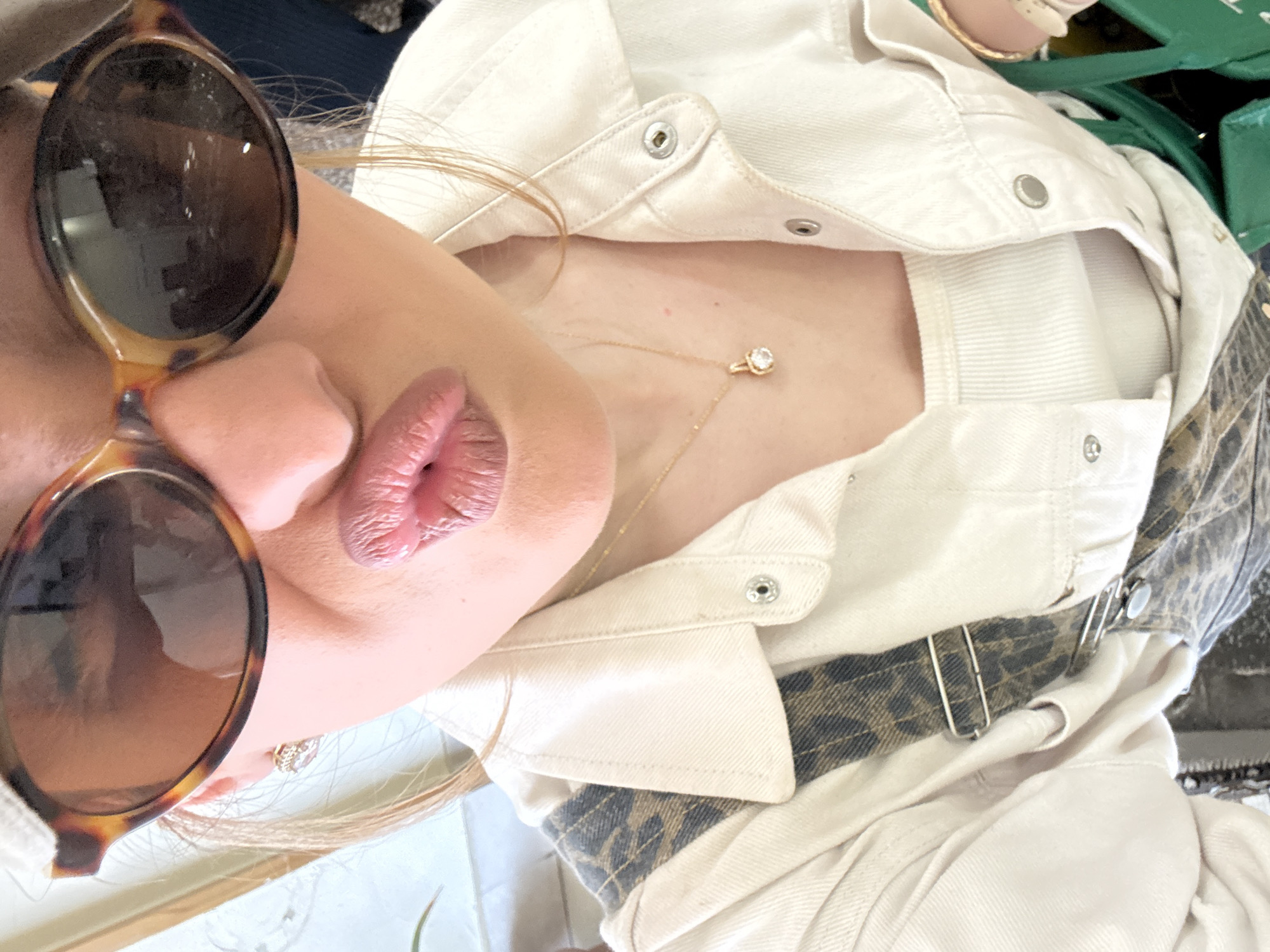A close-up shot presents a woman from an unusual angle, possibly a selfie or candid moment. She wears tortoiseshell sunglasses, and her lips are prominent, colored in a nude lipstick. A gold necklace with a diamond pendant rests on her fair skin. She is wearing a white denim jacket with snap closures and a white ribbed top underneath. Part of a leopard-print accessory is visible underneath the jacket. The background is blurred with hints of green and other textures. The location and city of Yerevan, Armenia are not discernible from the visual information available in the image.