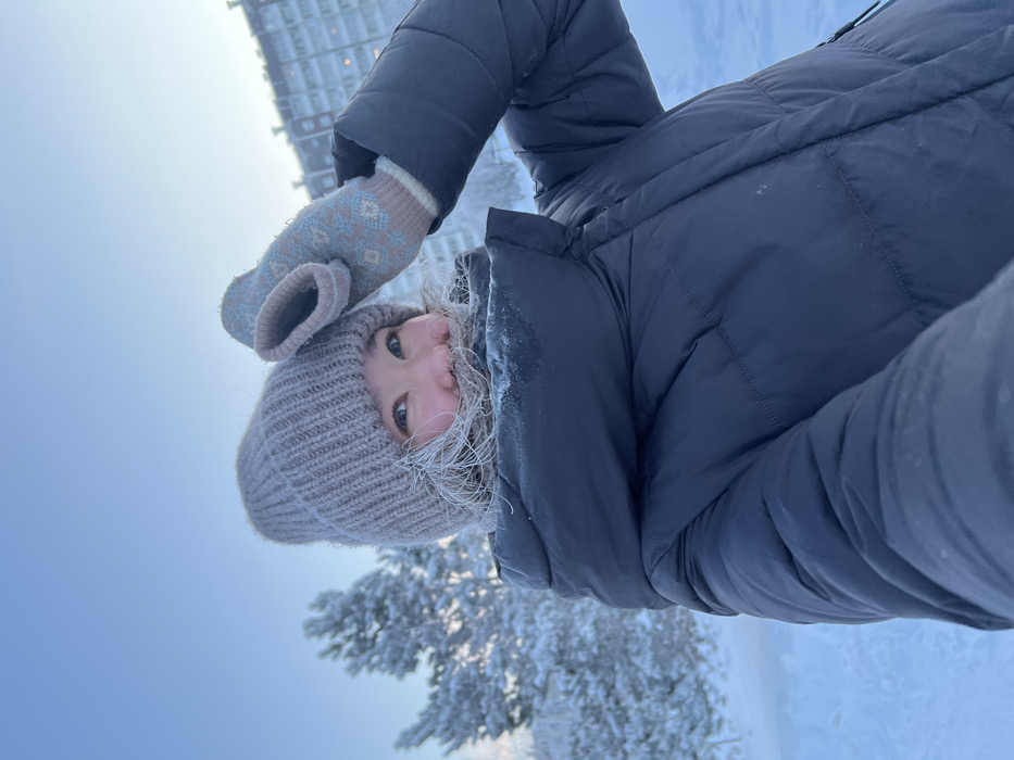 A single individual, identified as an adult female, is captured in a close-up, upward-angled selfie during extreme winter conditions. Her face is partially obscured by a light brown knit beanie and the high collar of a dark blue padded winter jacket. Her exposed hair around her face exhibits visible frost accumulation, as does the fabric of her jacket collar. Her eyes are open, and she displays a slight smile, revealing upper teeth. Her left hand, encased in a light brown mitten patterned with blue geometric designs, is raised near her head.
The immediate foreground is entirely covered in undisturbed snow. In the mid-ground to the left, snow-laden coniferous trees are visible. In the distant background to the right, a large, multi-story residential apartment building, light-colored with visible windows, stands against a clear, pale blue sky. The scene is illuminated by natural light, consistent with a bright winter day. The extensive snow, frosted elements, and heavy winter attire indicate severe cold. This scene is located in a park-like or residential area within the city of Krasnoyarsk, Russia.