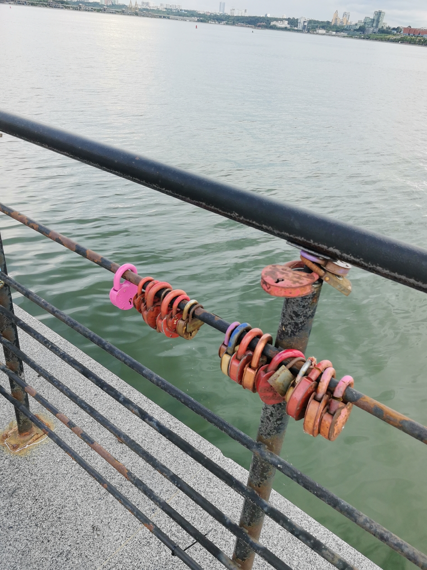 The image depicts a section of a railing overlooking a body of water, likely the Volga River, in Kazan, Russia. The railing is made of dark metal, with some sections showing rust. Attached to the railing are numerous love locks, predominantly red and pink, symbolizing commitment. In the background, the cityscape of Kazan is visible, characterized by buildings and structures along the shoreline. The water is calm, with a slight ripple effect. The lighting suggests it is daytime. There are no people or animals visible in the frame.