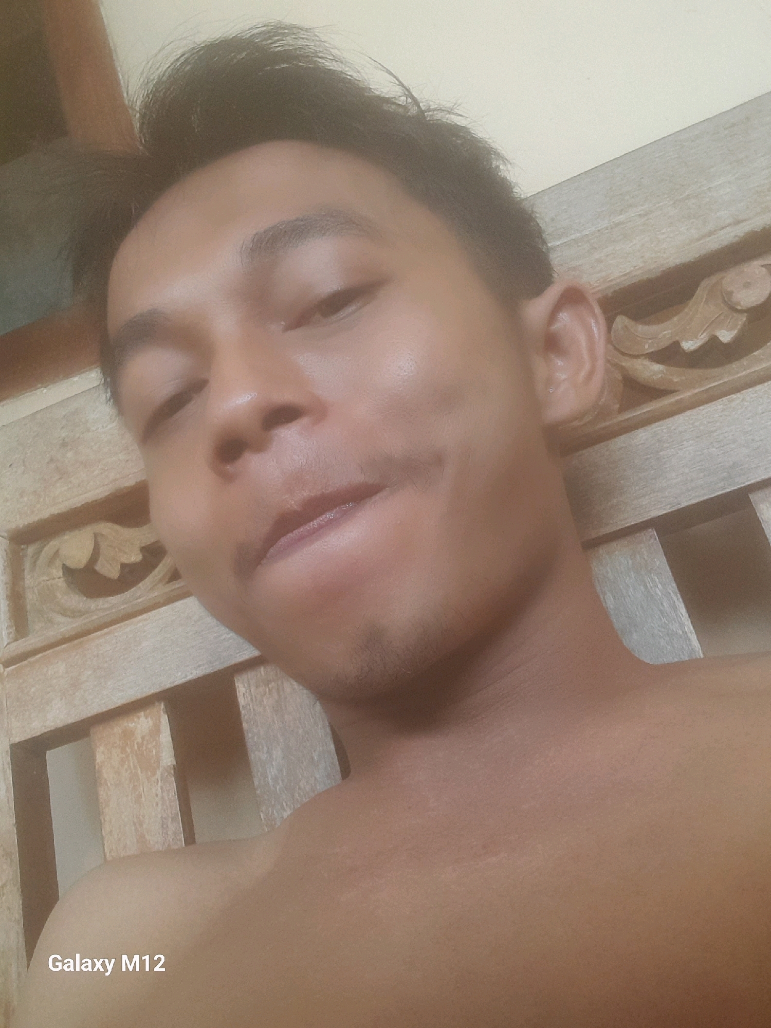 The image shows a close-up, selfie-style photograph of a young man's face and upper chest. He has dark hair, dark eyes, and fair to medium skin tone. He is looking slightly off-camera with his mouth partially open. The background appears to be a wooden structure, possibly a chair or furniture with carved details. The lighting suggests an indoor setting. The lower left corner of the image contains the text "Galaxy M12," indicating the device used to take the photo. The timestamp "03/12/23" is visible, suggesting the image was taken on March 12, 2023. There is no discernible context to place this image specifically within the city of Karawang, Indonesia.