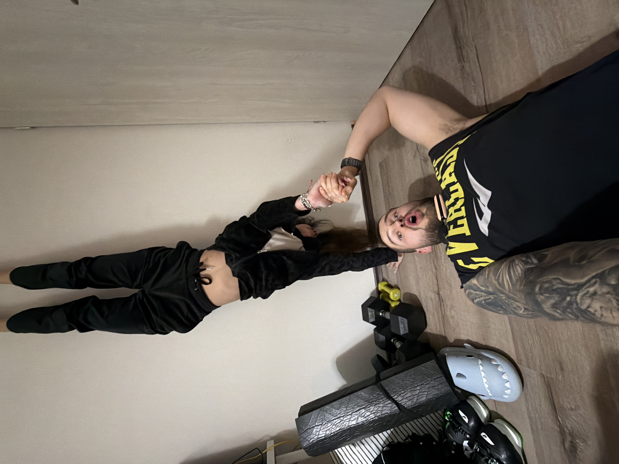 A person is lying on a wooden floor wearing a black Everlast t-shirt and shorts, showing a large tattoo on their arm. They are holding the hand of another person wearing a black outfit lying on a white wall. A set of dumbbells and exercise equipment are also on the floor. The location of this photo is likely indoors. There is no indication that this is in Sofia, Bulgaria.