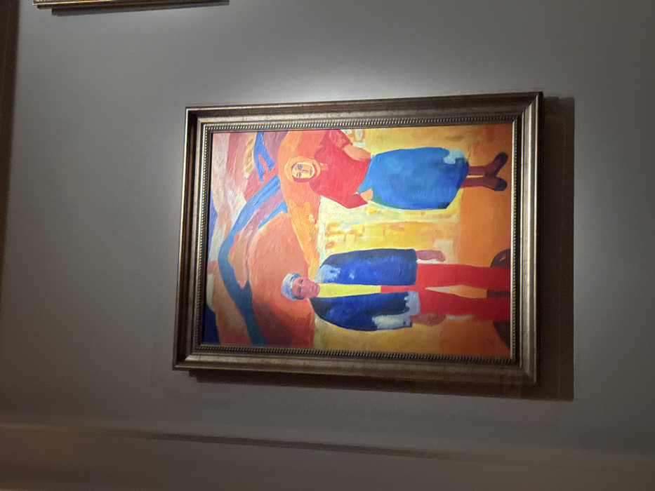 A framed painting hangs on a gray wall. The artwork features two stylized figures. One figure is dressed in a red shirt and blue skirt, appearing to float or levitate. Below, another figure is clad in a blue jacket and red pants, seeming to lie horizontally. The painting employs vibrant colors and abstract forms against a backdrop of orange and yellow hues. A simpler framed painting is partially visible above.