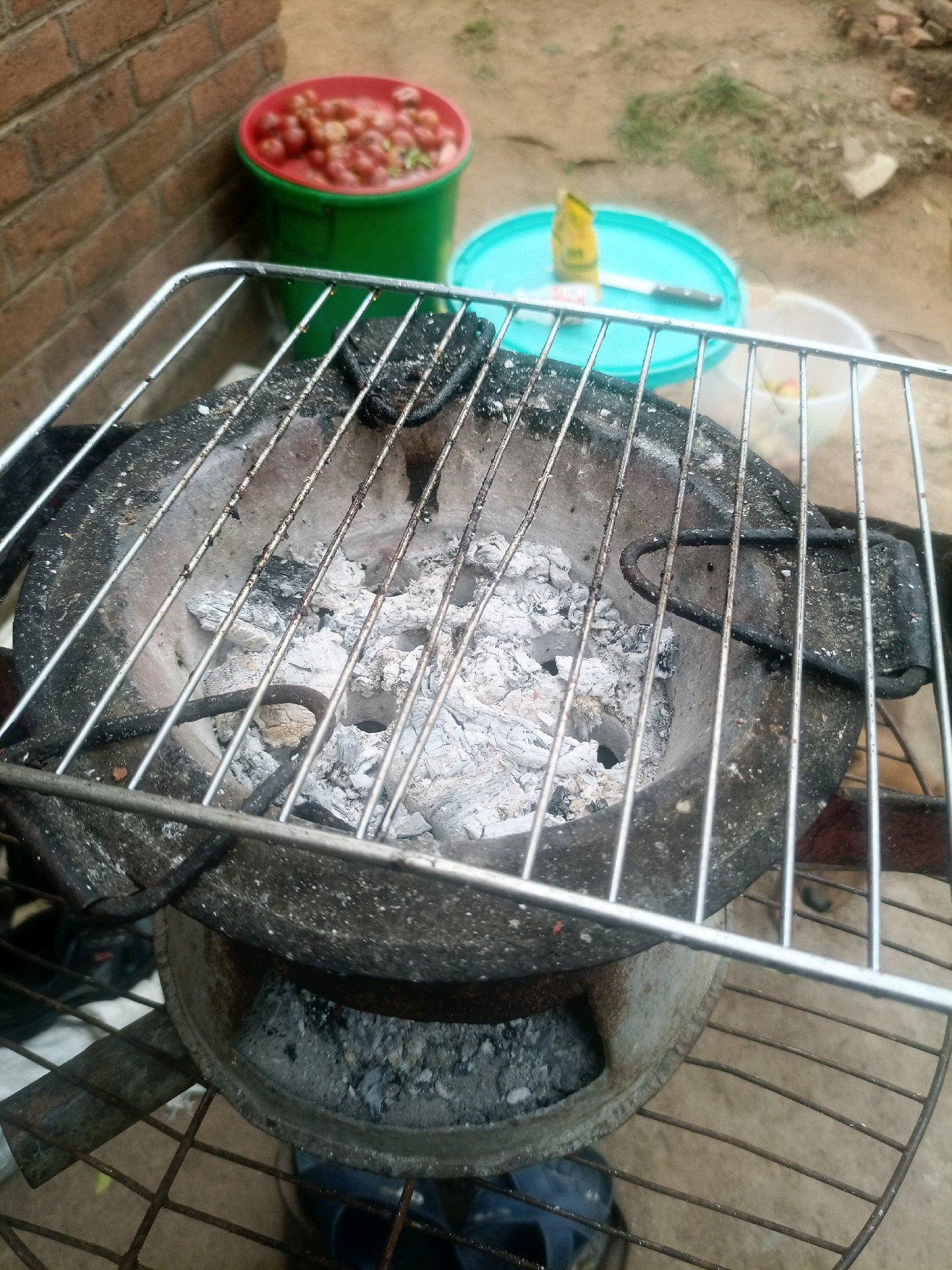 In Chiswe, Malawi, an outdoor scene depicts a charcoal grill with a metal grate placed on top. The grill contains white ash, indicating it was recently used. To the left of the grill, a red-rimmed green bucket is filled with small, red, round objects, possibly tomatoes or chilies, piled high. Behind the bucket, a brick wall is visible. To the right of the grill, a light blue lid is on the ground, with a yellow packet and a knife resting on it. Another white bucket, containing some items, is partially visible to the right. The ground is a mixture of dirt and dry grass. The overall impression is of an outdoor cooking or food preparation area.