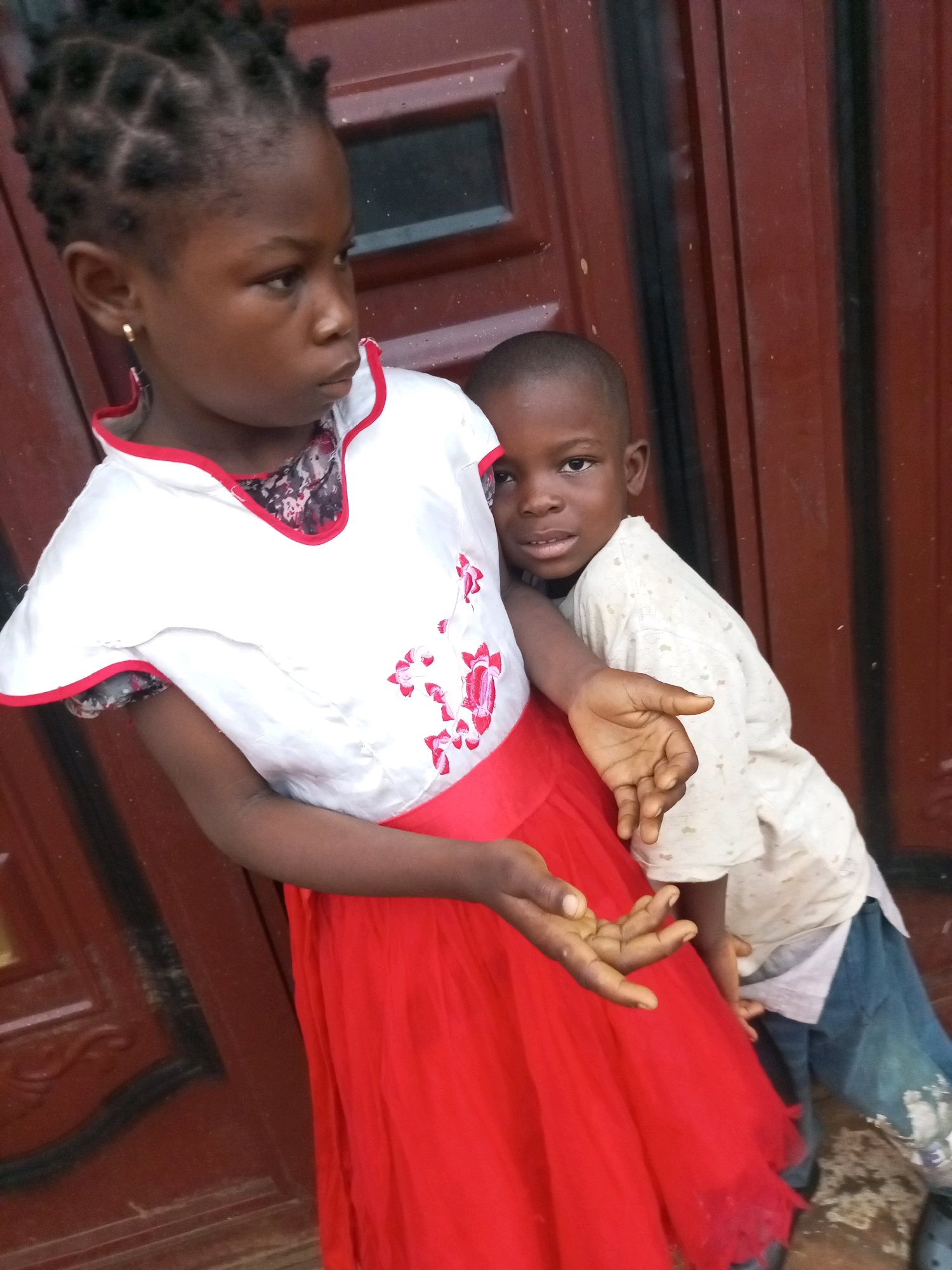 The image shows two young children, a girl and a boy, standing in front of a dark red door. The girl is in the foreground, wearing a white top with red trim and floral embroidery, and a long, flowing red skirt. Her hair is styled in small braids. The boy is behind her, leaning against her. He is wearing a light-colored, patterned t-shirt and blue jeans. Both children appear to be of African descent.
The girl is looking to her left with a serious expression, while the boy is looking towards the camera with a neutral expression. The girl's left hand is resting on her skirt, and the boy's right hand is reaching out, palm open. The background is a textured, dark red door with vertical black lines, suggesting an outdoor setting, possibly in a residential area. The lighting suggests it is daytime, but there are no specific clues about the weather. The overall scene depicts a moment of quiet interaction between the two children. There is no visible text in the image. The location context is provided as Tempure, Nigeria.