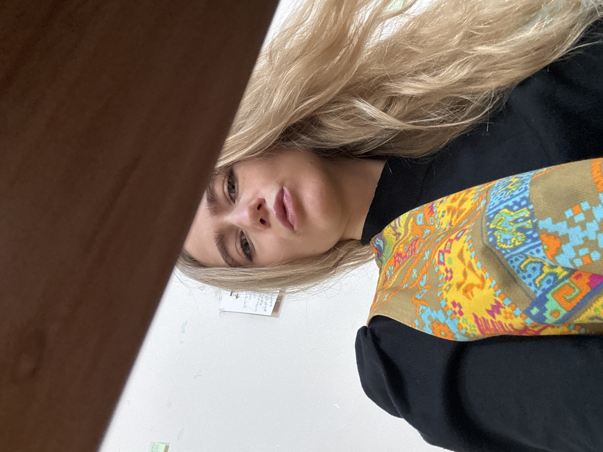 A low-angle, close-up photograph shows a woman indoors. She has long, wavy blonde hair and faces the camera directly with a neutral expression and slightly parted lips. She wears a black high-neck top, with a brightly patterned garment featuring yellow, orange, blue, green, and pink geometric designs on a tan background visible over her left shoulder. The upper portion of the frame is partially obscured by a dark, out-of-focus object, consistent with a wooden surface. The background consists of a plain, light-colored wall with several small sticky notes and a piece of paper containing handwritten Georgian script affixed to it. The specific location within the city of Unknown, Unknown, is not discernible from the image.