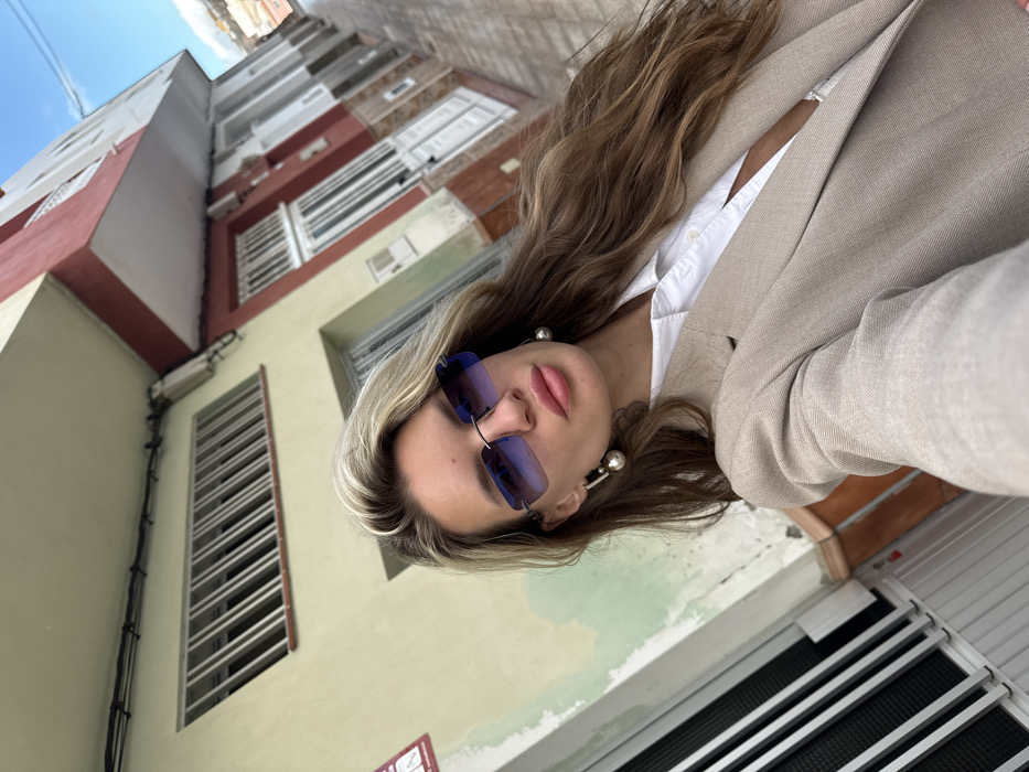 A female subject is depicted in a selfie, positioned centrally against an urban street scene in Arico, Spain. She has long, wavy blonde and light brown hair, wears rimless blue gradient sunglasses, large pearl-like drop earrings, a white collared shirt unbuttoned at the top, and a light beige blazer. She looks directly into the camera with a neutral expression. The background features two adjacent multi-story buildings. The building immediately behind the subject exhibits a faded yellow-green facade with a horizontal window secured by white vertical bars and a partially visible white sign with red text. To the subject's right, a multi-story building displays a reddish-brown lower section with white-framed windows and doors, and a lighter upper section featuring an overhang. The street pavement is visible in the lower portion of the frame, and a clear blue sky is visible in the upper right. The perspective is slightly angled upwards, capturing the street receding diagonally to the right.