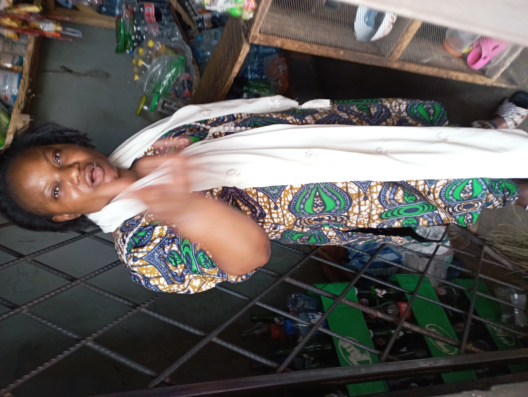 A woman stands behind a barred window in what appears to be a small store in Jos, Nigeria. She wears a patterned dress with green, blue, and brown designs, covered by a white shawl. Her hair is dark and pulled back. She is looking at the camera, gesturing with her right hand. Behind her, shelves are stocked with various items, including bottled drinks and packaged goods. Cases of bottled beverages are visible behind the barred window. The overall setting suggests a modest retail establishment in an urban environment.