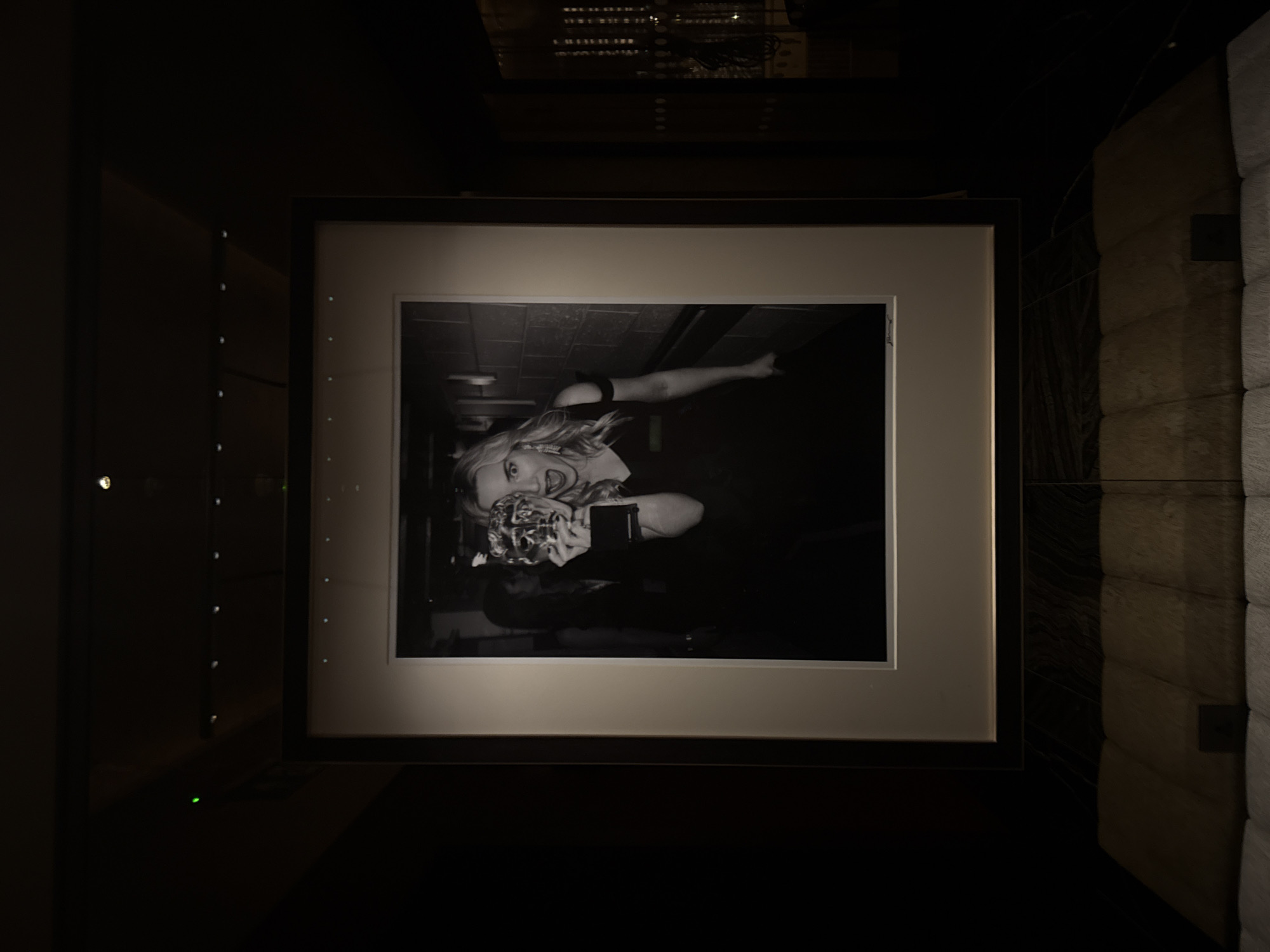 A framed black and white photograph hangs in a dimly lit room. The photograph features a blonde woman holding a statue, with a background suggesting an urban environment. The frame is dark, with a light-colored matte. The environment suggests a sophisticated or gallery-like setting. Due to the statue being a BAFTA award, the location is probably in London, United Kingdom.