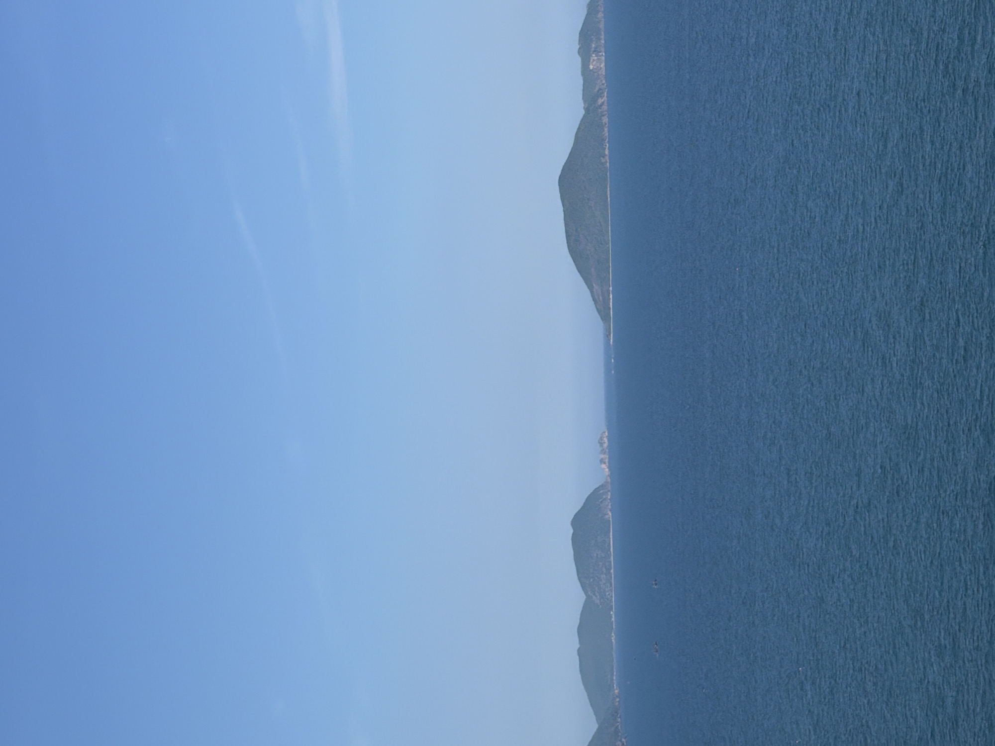 The image depicts a scenic view of the sea and islands under a clear blue sky. The horizon is visible, separating the water from the sky. Several lush green islands are scattered across the water, with some appearing closer and others more distant. The sea is a deep blue color, with subtle ripples on its surface, suggesting calm conditions. A few small boats are visible on the water, hinting at maritime activity. The scene is characteristic of coastal landscapes in the Nam Nha Trang Ward area of Vietnam, known for its beautiful beaches and islands.