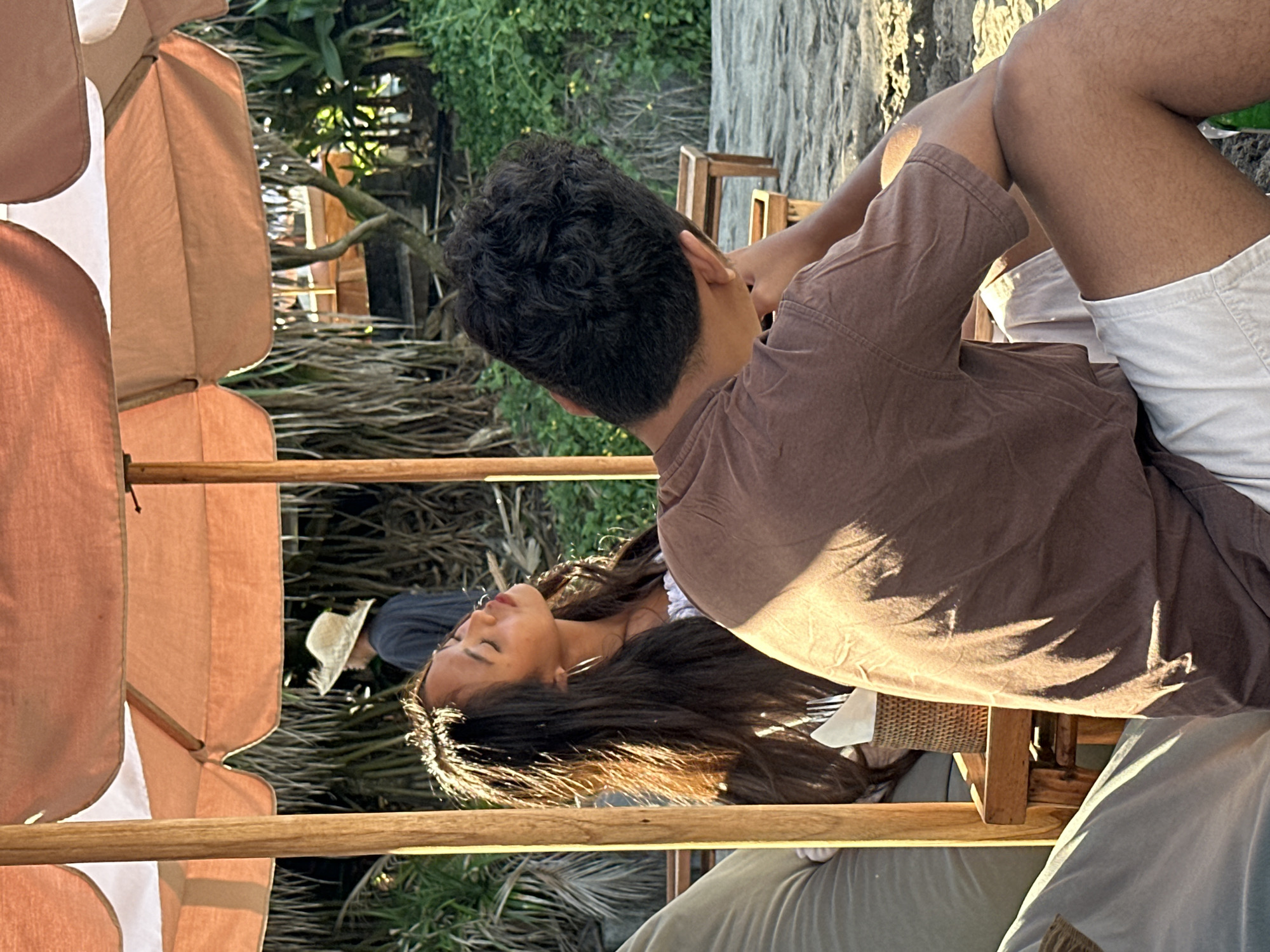 The image depicts a beach scene in North Kuta, Indonesia, under a large, peach-colored umbrella. In the foreground, a person with dark, curly hair is seen from the back, wearing a brown t-shirt and light-colored shorts, resting their head on their arm. To their left, a young woman with long, dark hair and hoop earrings is looking upwards with her eyes closed, bathed in sunlight, wearing a white top. Behind them, another person wearing a straw hat and dark clothing walks away. The background consists of lush green foliage and palm trees, with sandy ground in the immediate foreground. The overall atmosphere appears relaxed and summery.