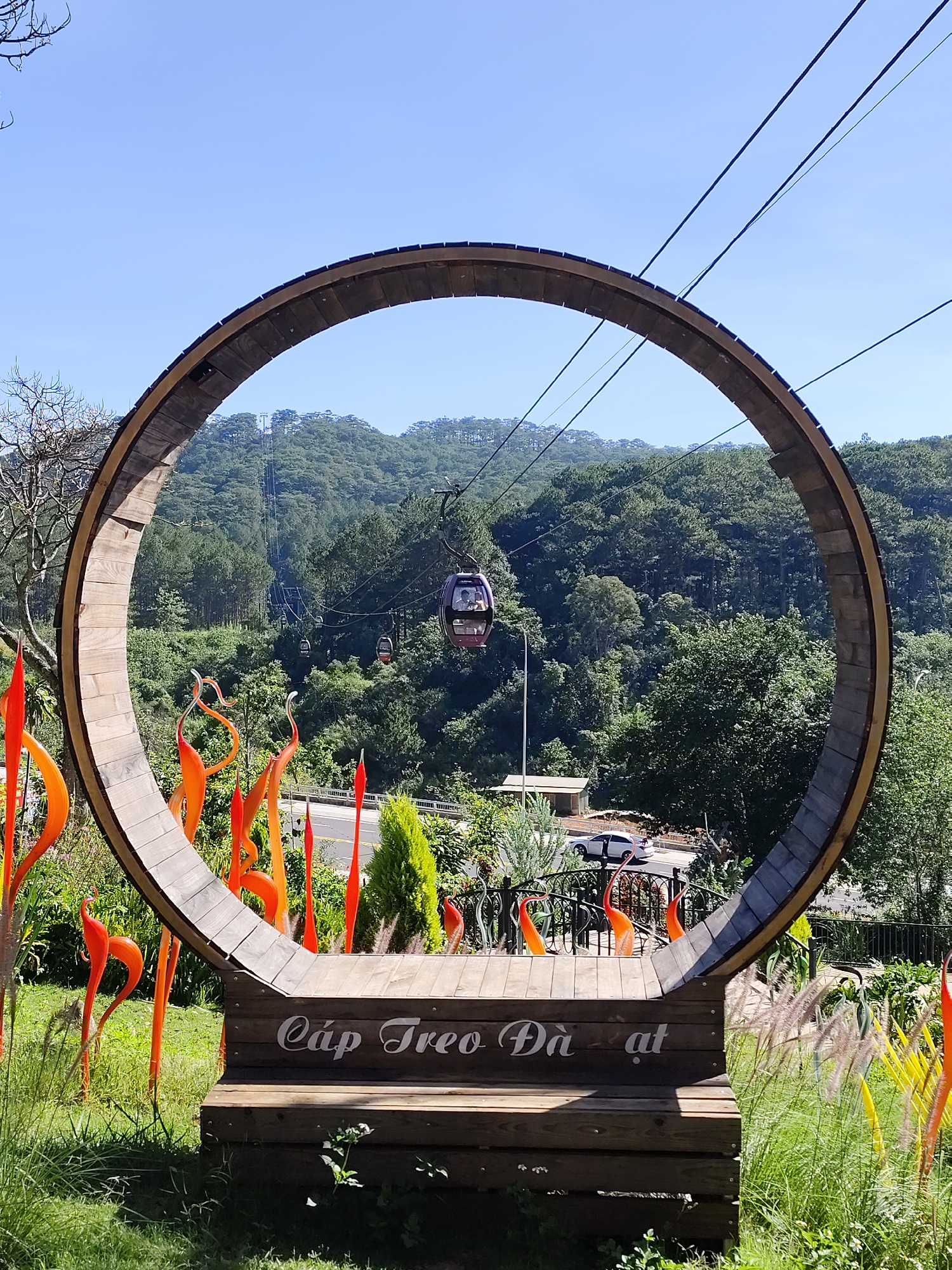 A medium outdoor shot shows a wooden circular structure sitting on a short, wooden bench with the words "Cáp Treo Đà Lạt" written in white script. The background is a lush, green landscape with pine trees, a roadway with vehicles, and a cable car moving up the mountain via suspension lines. The sky is clear and blue. In front of the circular wooden structure and bench are decorative glass pieces in shades of red, orange, and yellow, placed among green grass and plants. The wooden bench appears to be made of stacked wooden planks. The scene is likely in Da Lat, Vietnam, suggested by the words on the bench which translate to "Da Lat Cable Car."