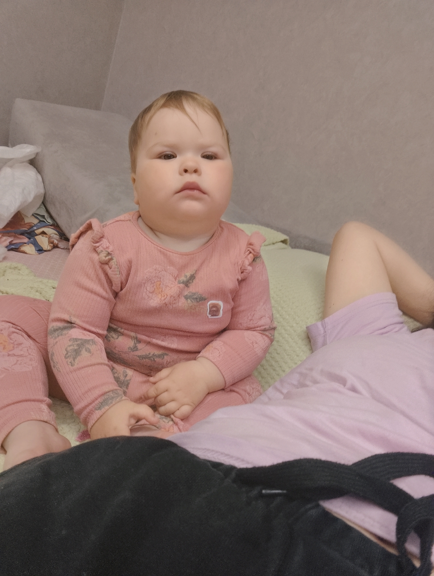 The image depicts an indoor scene in Chaikovsky, Russia, featuring an infant and partial view of an adult on a bed or large cushioned surface.
A light-skinned infant, appearing to be between six and eighteen months old, sits upright in the center of the frame, facing directly towards the viewer. The infant has light brown hair, dark eyes, and a neutral or slightly serious expression. They are dressed in a long-sleeved, ribbed pink top with ruffled shoulders and a faded floral or botanical pattern in lighter tones across the chest and sleeves. Matching ribbed pink pants with a similar pattern are worn, and the infant's bare feet are visible. The infant's hands are clasped or resting on their lap.
To the right of the infant, the lower left leg and upper left thigh of an adult are visible, suggesting the adult is reclining. The adult's leg is covered by a light purple or lavender fabric, likely shorts or pajamas. The foreground is dominated by a crumpled mass of dark, possibly black, fabric, which includes a visible drawstring or strap, obscuring the adult's lower body and part of the infant's lower legs.
The subjects are situated on a light green, textured blanket or throw, which covers a larger, light-colored bed or cushion. In the background, behind the infant, is a large, light grey or off-white pillow or cushion. To the left, a crumpled white tissue or cloth is visible. The wall behind the bed is a plain, light grey or beige color with a subtle texture.