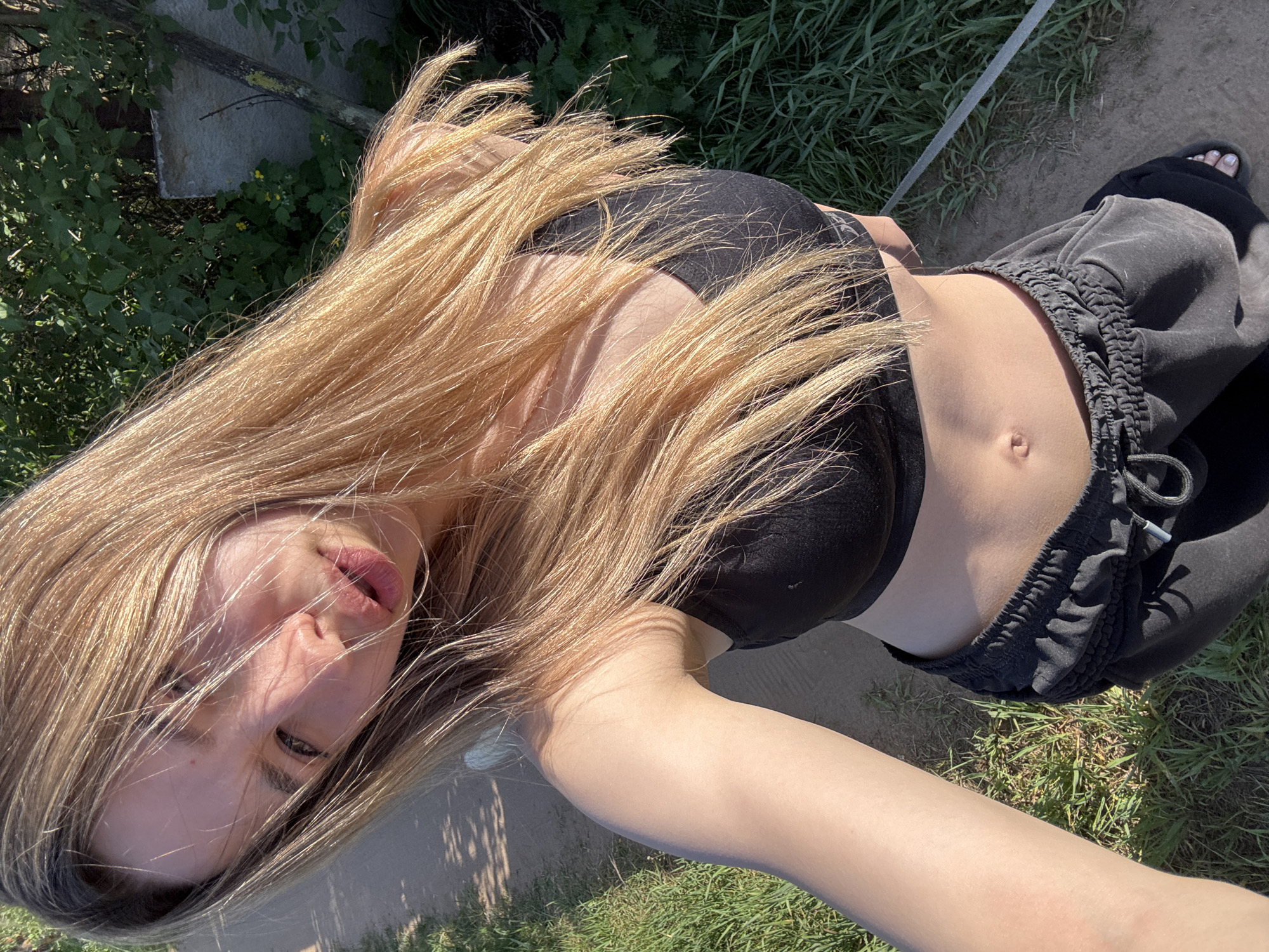 A woman is posing for a selfie outdoors. She has long blonde hair and is wearing a black sports bra and grey sweatpants. The woman is standing on grass and dirt. In the background, there is a concrete slab and some greenery. There is no information to indicate that this image was taken in the city of Noviy, Russia.