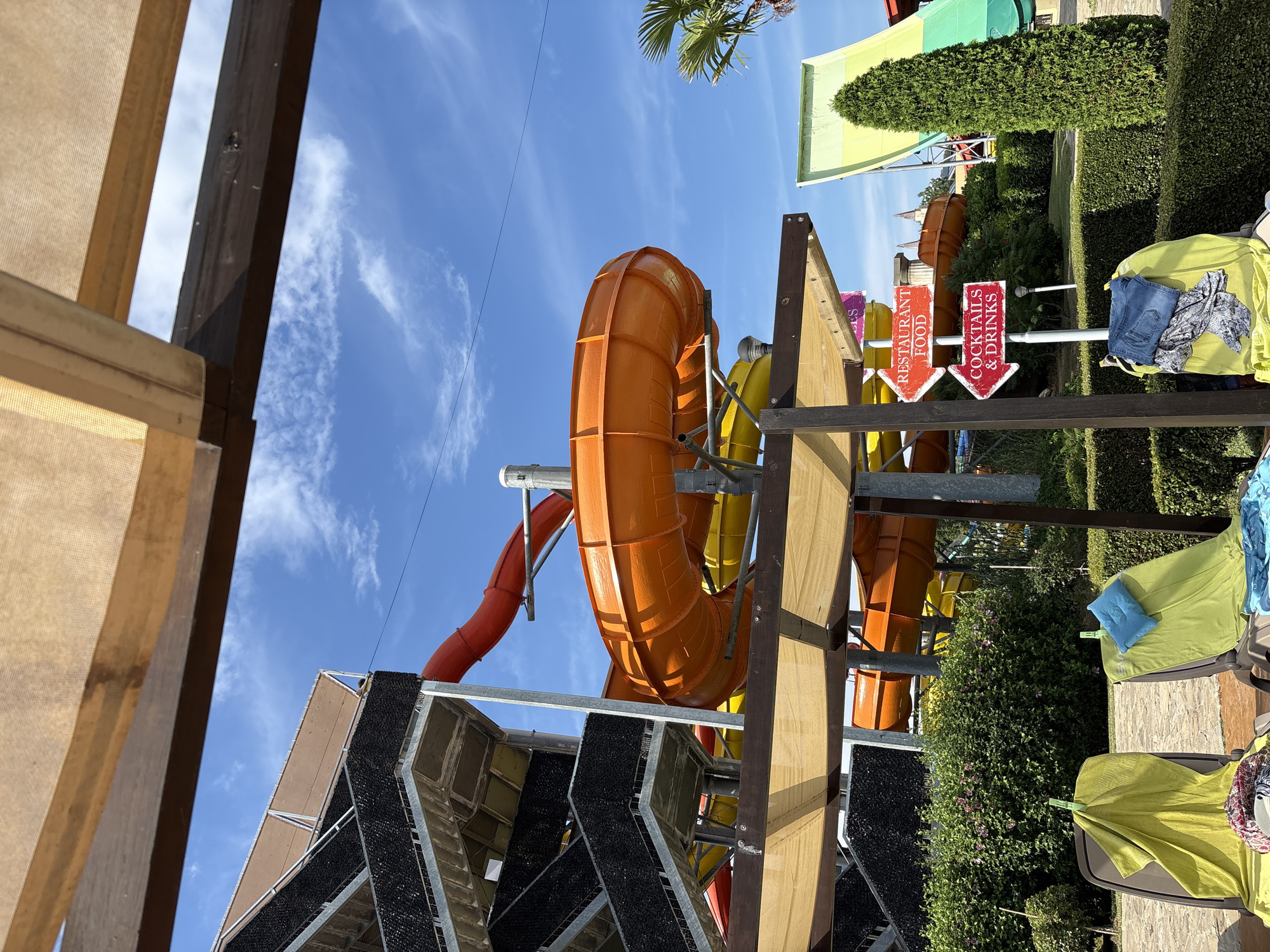 The image captures a sunny day at a water park, likely in Nesebar, Bulgaria. The primary subjects are several brightly colored water slides, one prominent orange slide with a curved section, and a section of a red slide visible to the left. Yellow slides are also present further back. The water park features lush green hedges and manicured bushes, suggesting a well-maintained outdoor recreational area. In the foreground, part of a wooden structure with a beige fabric canopy is visible, offering shade. On lounge chairs to the right, lime green towels and a blue pillow are laid out, alongside some folded clothing, including blue jeans.
The activity depicted is relaxation and the anticipation of water park fun. The clear blue sky with wispy white clouds indicates fair weather and a pleasant time of day, likely late morning or afternoon, given the bright sunlight casting shadows. Location cues include the characteristic Mediterranean architecture visible in the background structures and the style of the water slides. There are several signs with text: two red arrow signs pointing left and right, one reading "RESTAURANT FOOD" and the other "COCKTAILS & DRINKS". Other signs with illegible text are also present further back. The overall mood is one of leisure and enjoyment, characteristic of a vacation setting.