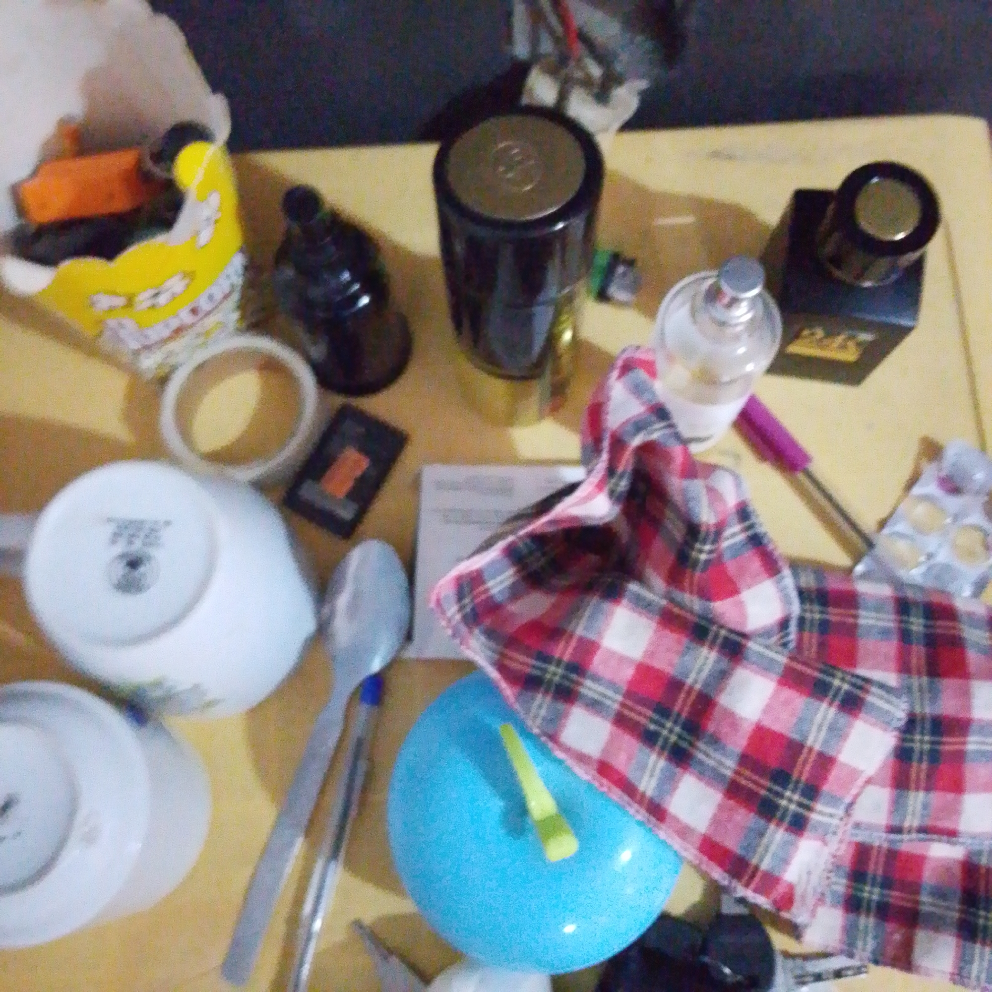 The image depicts a cluttered tabletop scene, likely indoors, with various objects scattered across a light-colored surface. In the foreground, a blue sphere, possibly a balloon or a stress ball, is partially covered by a red and white plaid cloth. To the left of the blue sphere, two silver spoons rest side-by-side, with one exhibiting a blue tip. Behind the spoons and to the left, a white ceramic cup is visible, its interior dark. Further left, a white bowl or saucer sits with its underside facing upward, revealing some text and a logo. At the far left, a paper bag with yellow and orange elements, appearing to hold small items, is present.
Dominating the mid-ground are several bottles and containers. A dark, cylindrical bottle with a gold lid stands prominently. To its left, a smaller, dark bottle with a reflective surface is partially visible. Next to the dark cylindrical bottle, a clear glass bottle with a silver cap contains a light-colored liquid. A pink and purple pen or stylus lies diagonally across the plaid cloth, near the clear bottle.
In the background, the scene is darker and less distinct. A small, dark rectangular object with an orange element is situated between the dark bottles and the spoons. A few other unidentifiable objects are present in the background, blending into the shadows. There are no people visible in the image. The overall impression is one of disarray, with personal items or craft supplies potentially laid out for use or storage.
The location in Jalingo, Nigeria, is not deducible from the visual information presented in the image.