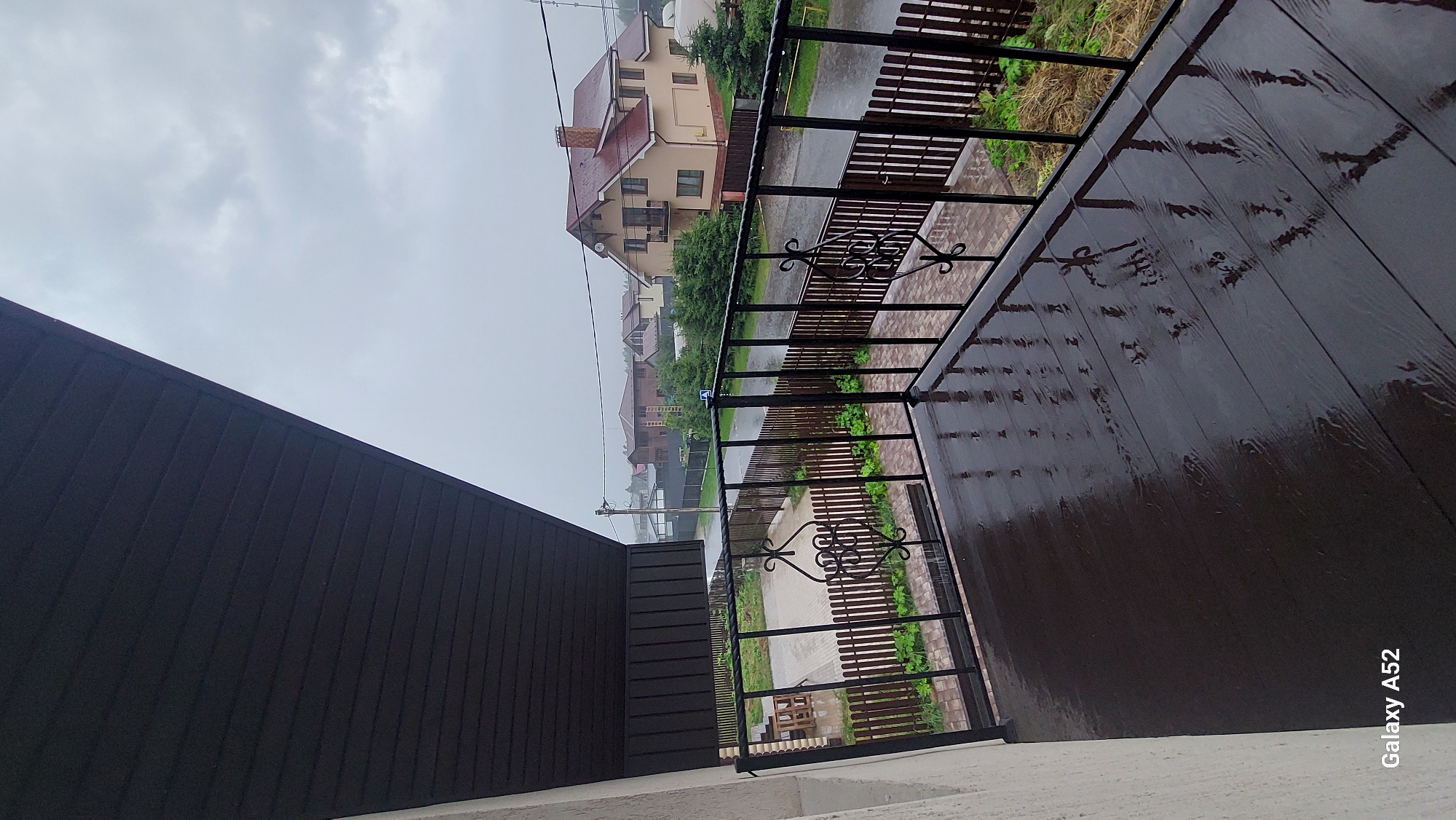 The image shows a wet, wooden balcony overlooking a street in a residential area during a rain shower. The balcony floor is dark brown and reflects the sky and surrounding objects due to the wetness. A black metal railing with ornate scrolling decorates the edge of the balcony. Beyond the railing, a wooden fence encloses a yard with some patchy grass and dry vegetation. A paved driveway is visible. Across the street, houses with various roof styles and colors are nestled among green trees. The sky is overcast and gray, indicating rainy weather. The scene appears to be located in the city of Dolche Vita KP, Russia, as suggested by the device watermark in the bottom left corner, "Galaxy A52". There are no people visible in the image.