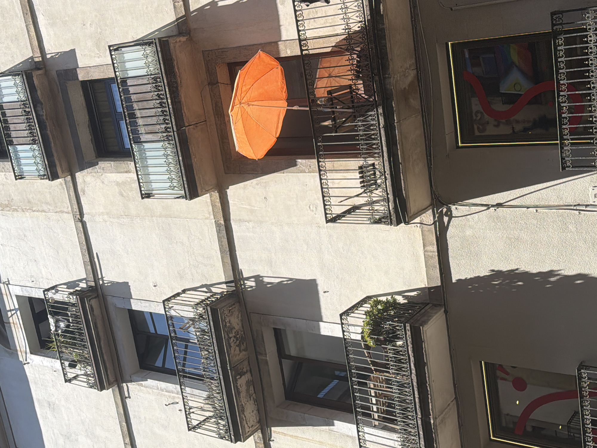 An eye-level, tilted shot shows an exterior building wall in Barcelona, Spain. The exterior is light gray, with several balconies. Each balcony has a black iron rail. One of the balconies has an orange umbrella hanging in the middle. Each balcony is adjacent to a window. Some windows contain art or plants.