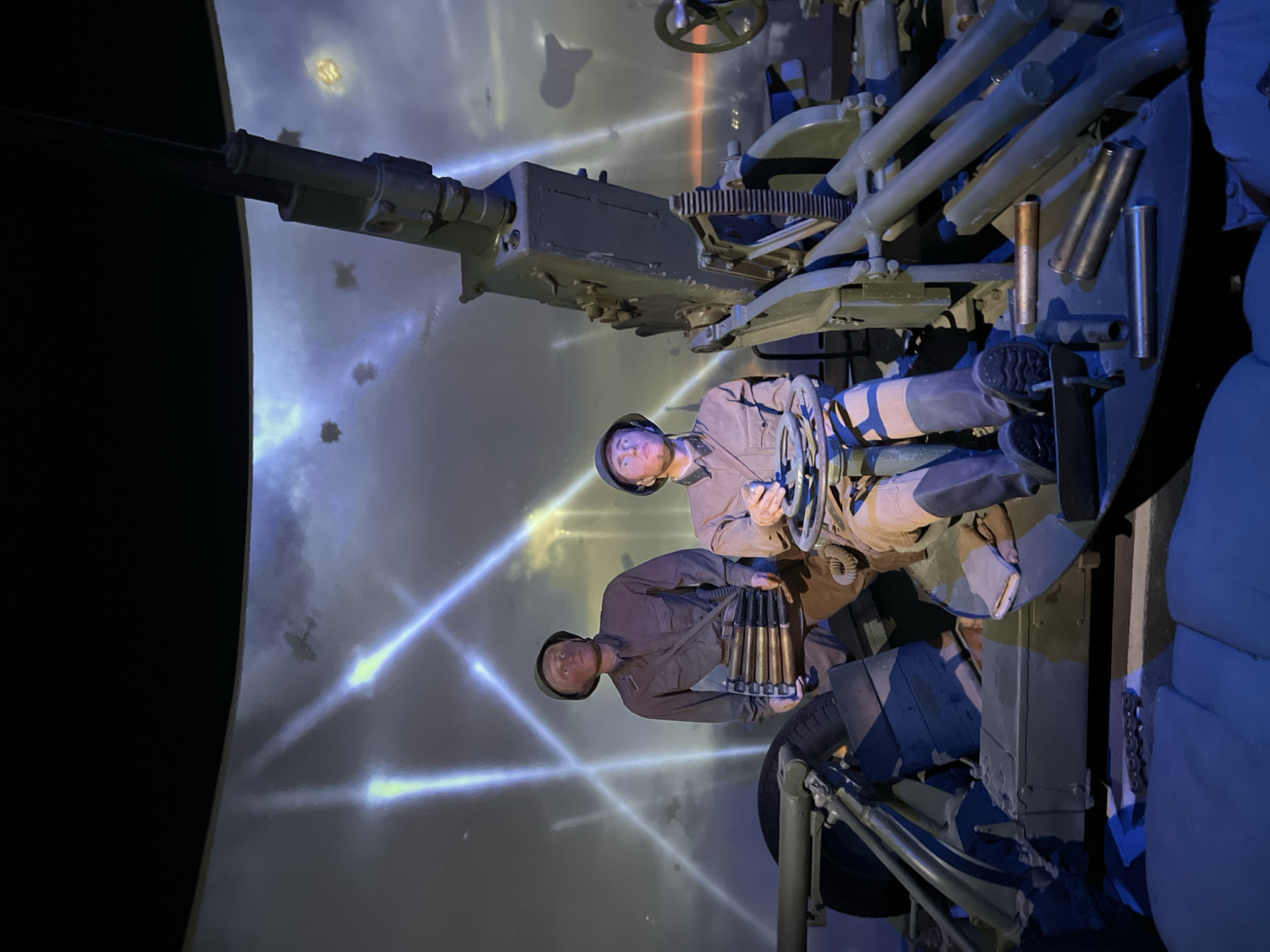 The image depicts a diorama featuring two mannequins dressed as soldiers operating an anti-aircraft gun. The mannequins are positioned within the gun emplacement, with one manning the controls and the other holding a belt of ammunition. The anti-aircraft gun itself is a prominent feature, painted in olive drab and appearing to be made of metal. In the background, a projected image displays a night sky with searchlights crisscrossing, and silhouettes of airplanes. The overall scene suggests a wartime setting, likely representing a defensive position. The question about the location in the city of Vetka Gertsena, Russia, is not supported by any visual information in the image itself.