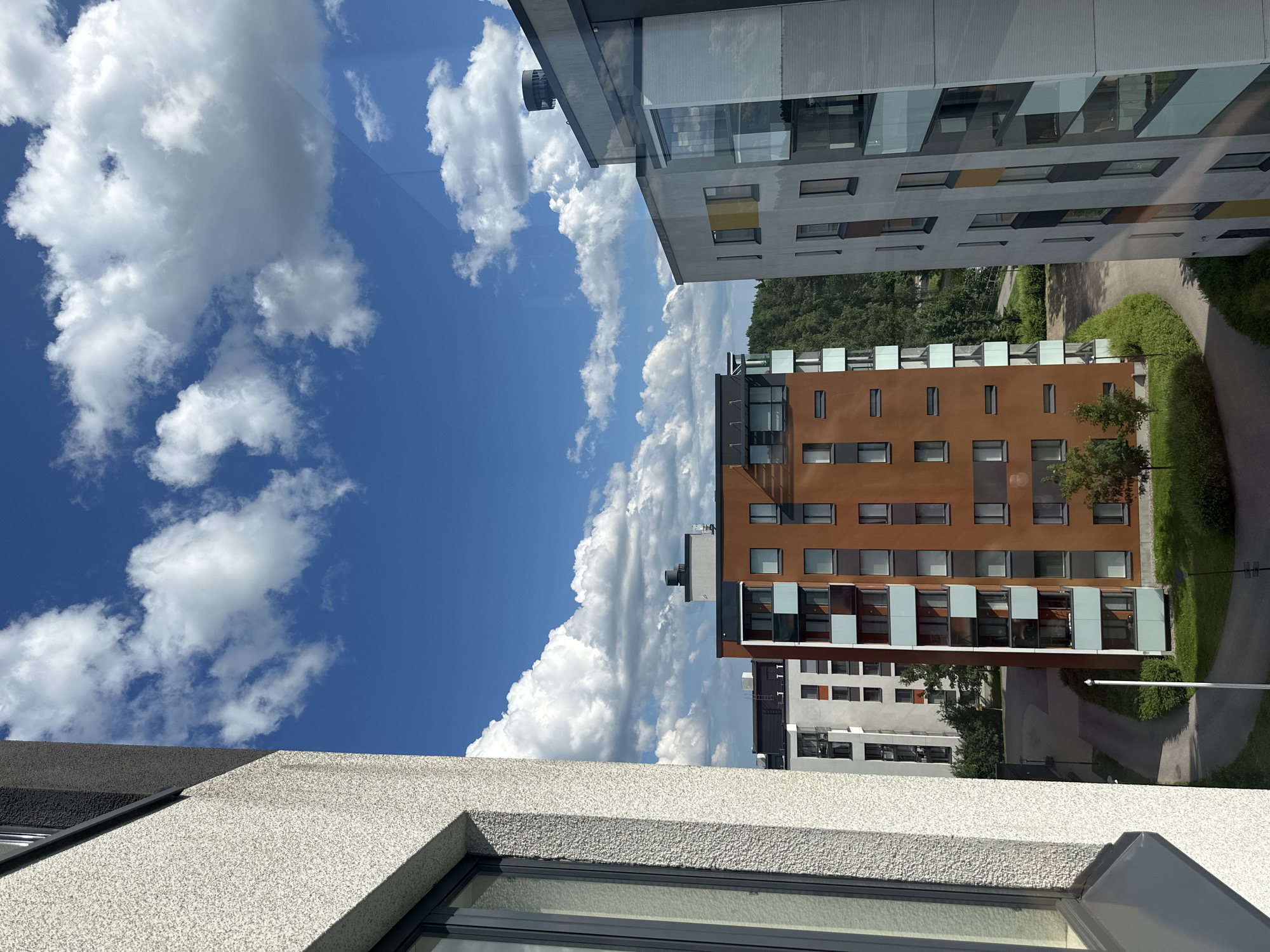 The image shows a view of a modern apartment complex under a bright blue sky with fluffy white clouds. The perspective is from a window, looking out and slightly upwards. In the foreground, the textured white facade of a building is visible, with a dark grey angled roofline and a window.
Dominating the mid-ground are two apartment buildings. The one closer to the camera is a striking orange-brown color with many windows and balconies that have glass panels. Behind it, a lighter colored building with a grey facade and rows of windows can be seen. The architecture suggests a contemporary urban or suburban setting. A paved pathway and green grassy areas are visible between the buildings. Trees with lush green foliage form a backdrop behind the complex.
The lighting and the clear blue sky with scattered clouds indicate it is daytime, likely sunny and pleasant weather. There is no visible text in the image. The scene evokes a sense of calm and urban living.