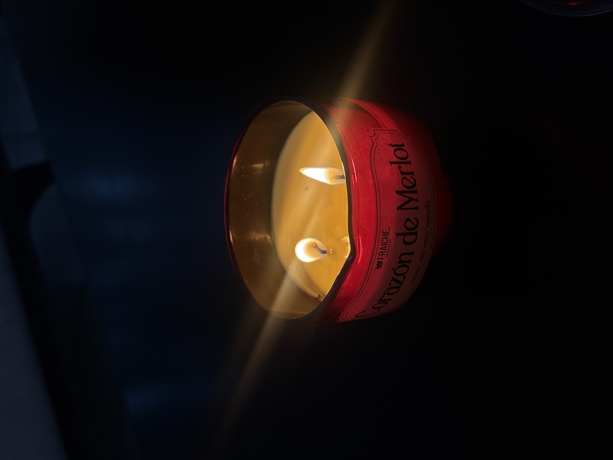 A close-up, low-angle shot shows a burning candle inside a red metallic container. The label reads "Corazón de Merlot" with additional text in Spanish. Two flames are visible within the melted wax. The background is dark, with a blurred blueish element to the left. The location is not discernible from the image.