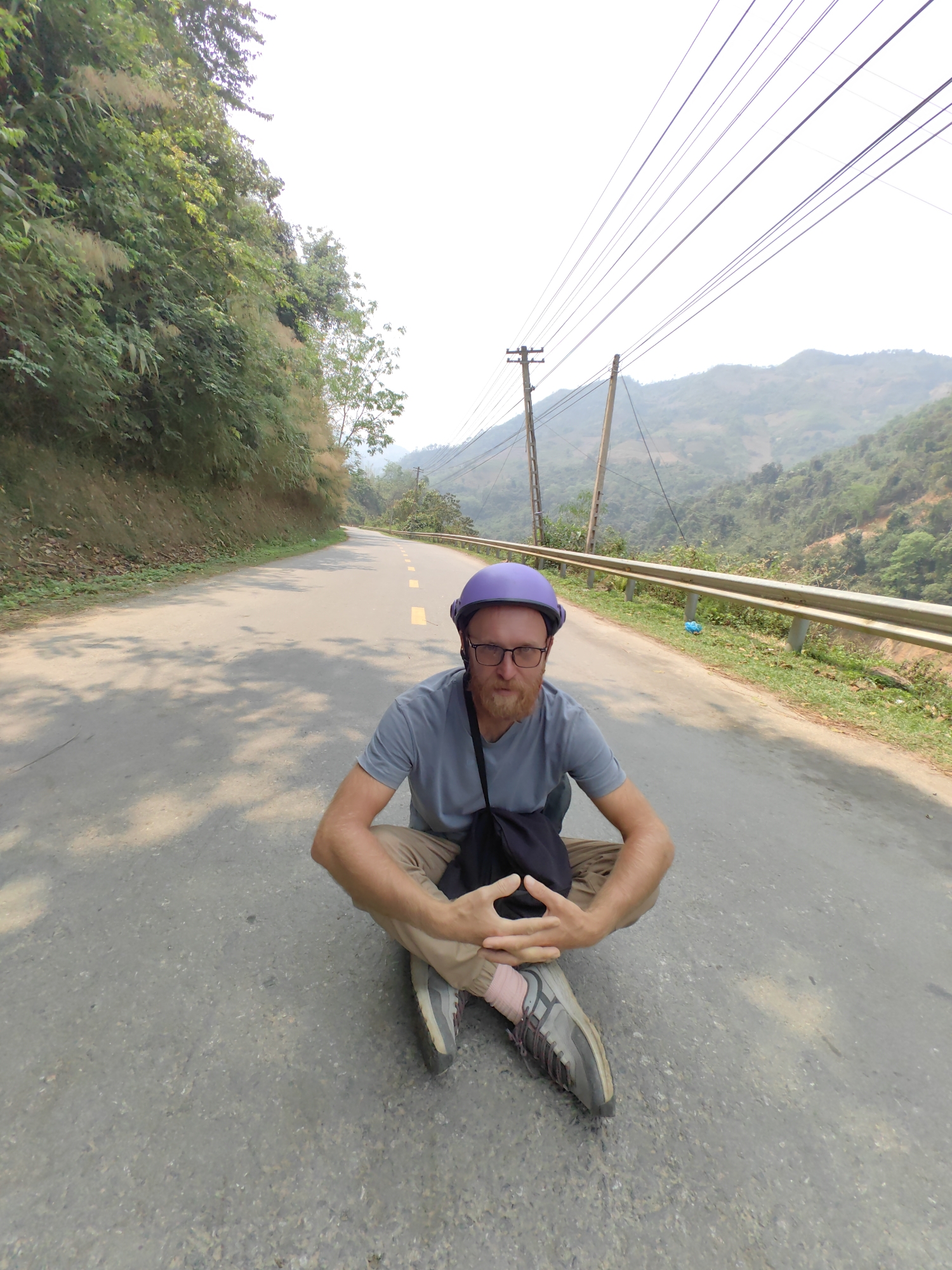 A man wearing a purple helmet, glasses, a gray t-shirt, and khaki pants is sitting cross-legged on a paved road. His hands are clasped together in front of him. He is wearing gray and purple shoes and has a black bag slung across his shoulder. The road is marked with yellow dashed lines down the center and has a metal guardrail on the right side. Lush green vegetation covers the hillside to the left of the road. Mountains are visible in the distance under a hazy sky. Utility poles with multiple wires run along the right side of the road. The location is along a rural road in the city of Bảo Lạc, Vietnam.