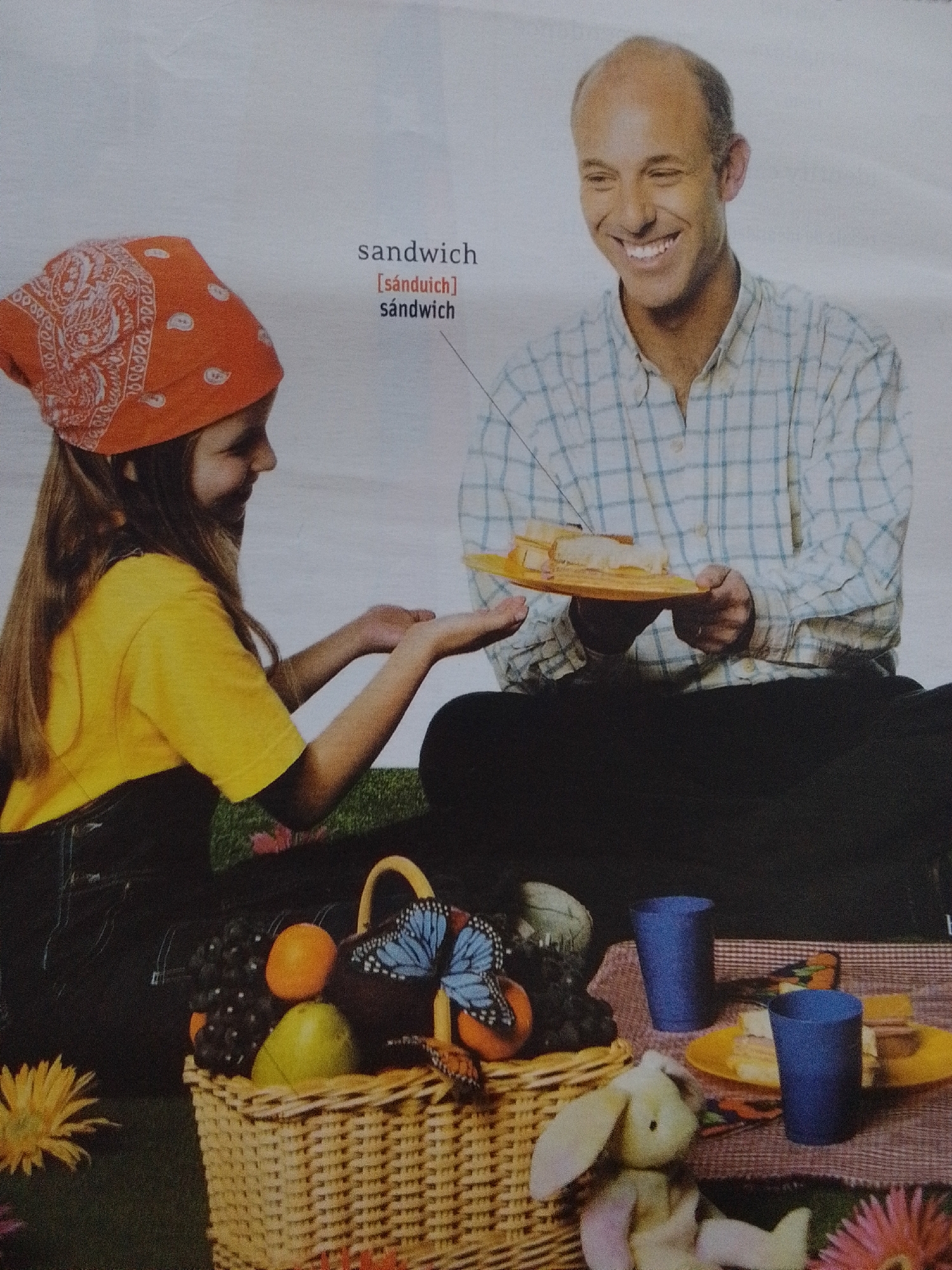 A man and a young girl are having a picnic. The man, bald and smiling, wears a blue and white checkered shirt and is offering a plate of sandwiches to the girl. The girl, with long brown hair and wearing a red bandana, a yellow t-shirt, and dark overalls, is reaching out her hands to receive the plate. They are sitting on the grass, with a picnic basket filled with fruit and decorated with butterflies beside them. Two blue cups and a plate with more sandwiches are on a red and white checkered blanket. The scene is set in a natural environment, possibly a park or backyard, with flowers visible in the foreground. There is no information in the image to suggest a location in La Tagua, Venezuela.