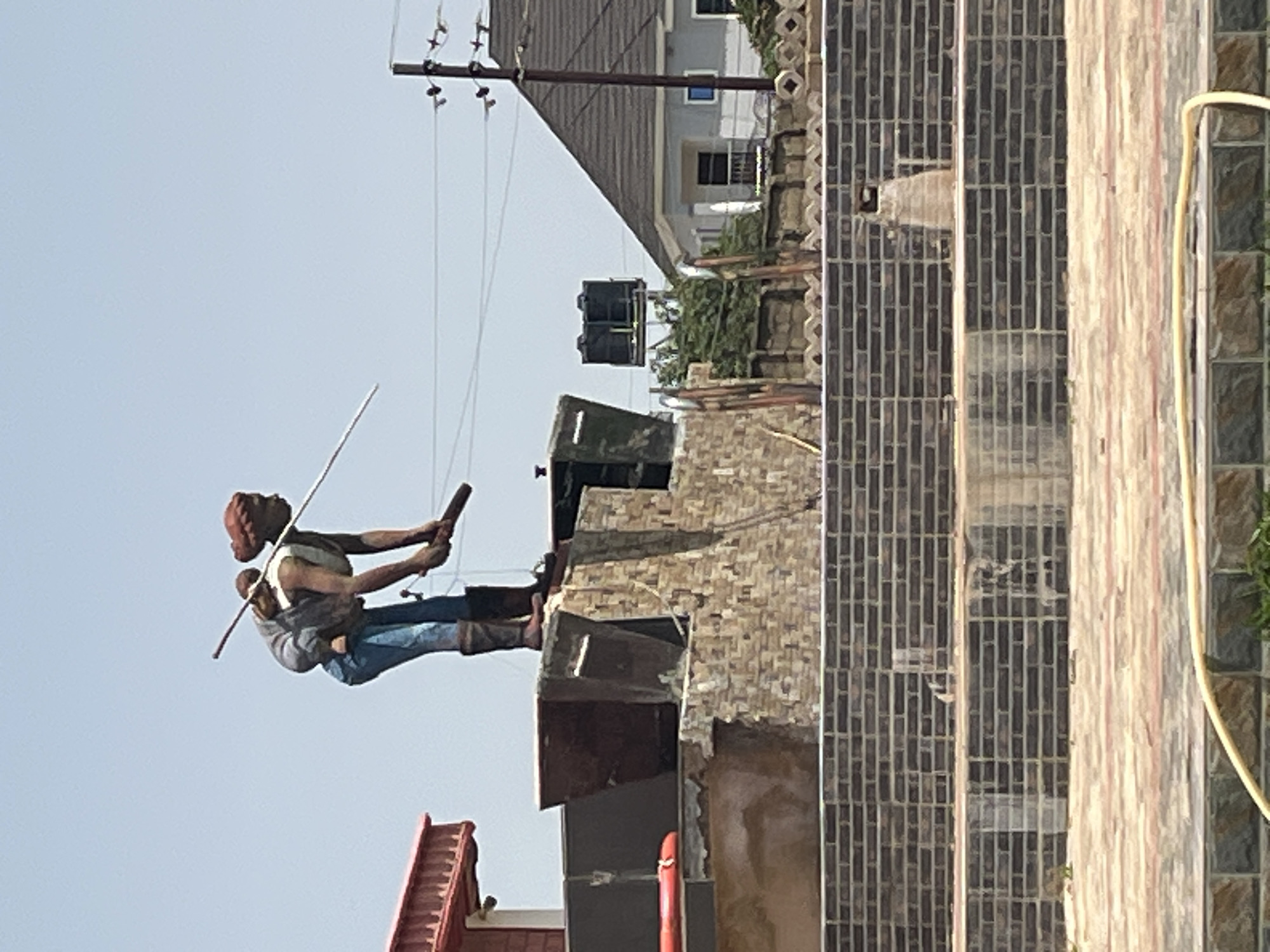 This image depicts a statue of a woman carrying a child on her back, holding a long stick. The statue is positioned on top of a multi-tiered, brick-faced structure, possibly a fountain or monument. In the background, a red-roofed building is visible to the left, while to the right, a modern house with a dark-tiled roof and a blue window is situated near utility poles and overhead wires. Two large black water tanks are also visible in the background. The foreground shows a tiled walkway. The scene appears to be outdoors in a city, with a clear sky. This statue is located at the junction of University of Jos Road and Murtala Muhammed Way in Jos, Nigeria.