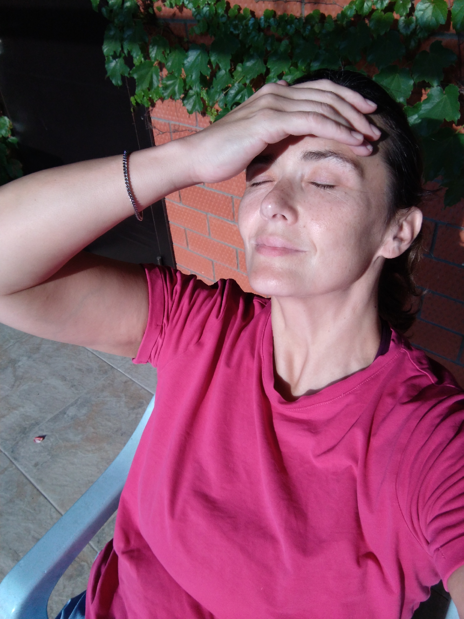 A woman sits on a light blue chair outdoors, with her eyes closed and hand on her forehead as if shielding herself from the sun. She wears a bright pink t-shirt. Behind her, a brick wall is covered with dense green ivy. Sunlight casts a shadow across her face, highlighting her freckles and a bracelet on her wrist. The scene suggests a warm, sunny day in Vinogradny, Russia, possibly during the daytime. The woman's expression conveys a sense of relief or discomfort from the bright sun.