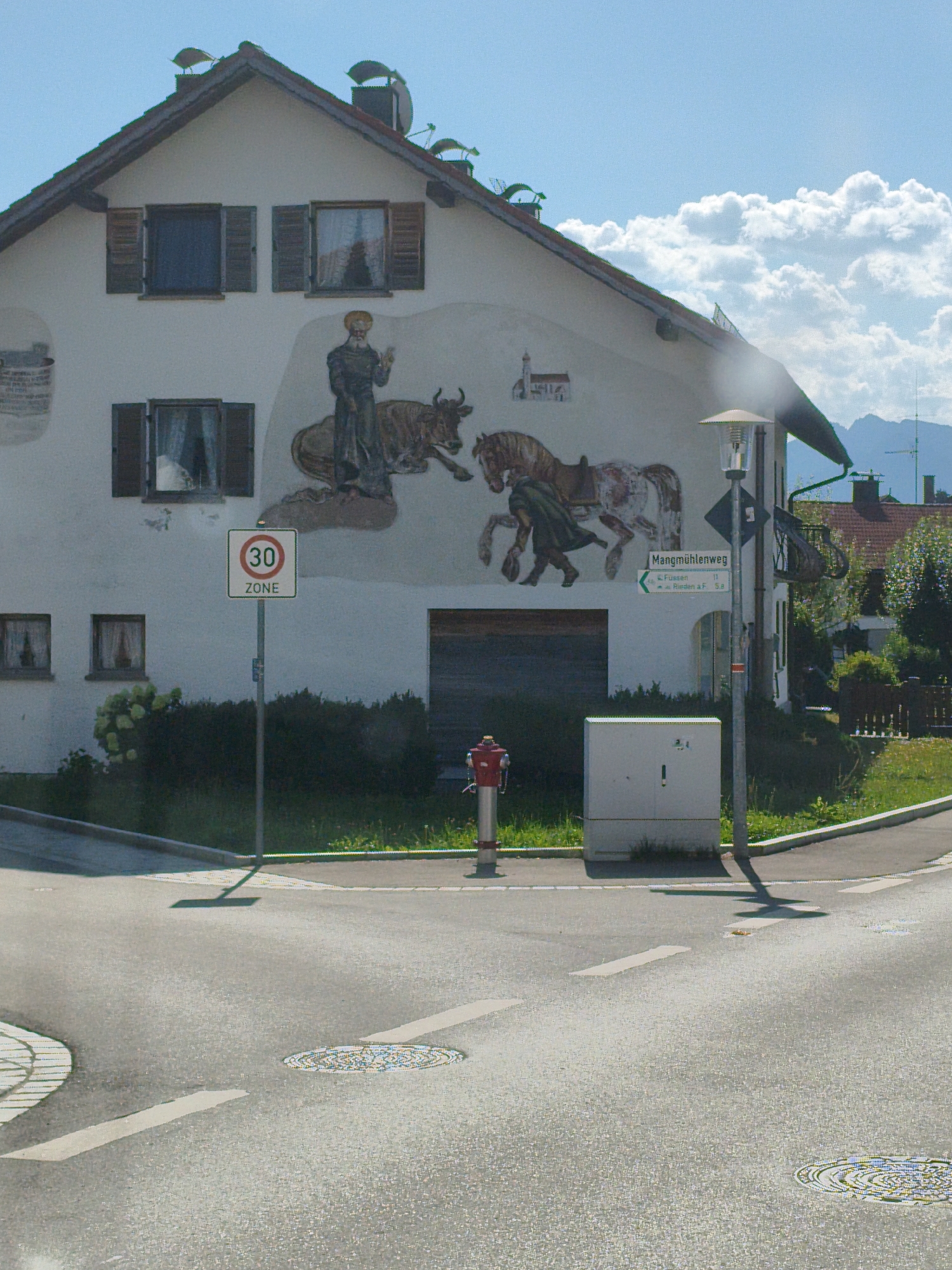 This media file captures a sunlit street scene in Roßhaupten, Germany. The main subject is a building adorned with a mural depicting a figure resembling a saint with a halo, leading a bull, and two horses with a figure in a green cloak. The mural is painted on the white facade of a two-story house with several windows and wooden shutters.
In the foreground, a road junction is visible with white markings and manhole covers. A speed limit sign indicating "30 ZONE" is prominently displayed. Adjacent to the sign is a lamppost and a directional sign pointing to "Mangmühlenweg" with distances to "Füssen" (11 km) and "Roßhaupten" (0.5 km).
Other objects in the scene include a red fire hydrant and a utility box. Green hedges and manicured grass border the street. The sky is blue with scattered white clouds, suggesting a pleasant day. The presence of mountains in the background indicates a mountainous or hilly region. The overall atmosphere is peaceful and rural. No specific activity or event is taking place, it appears to be a static view of the location.