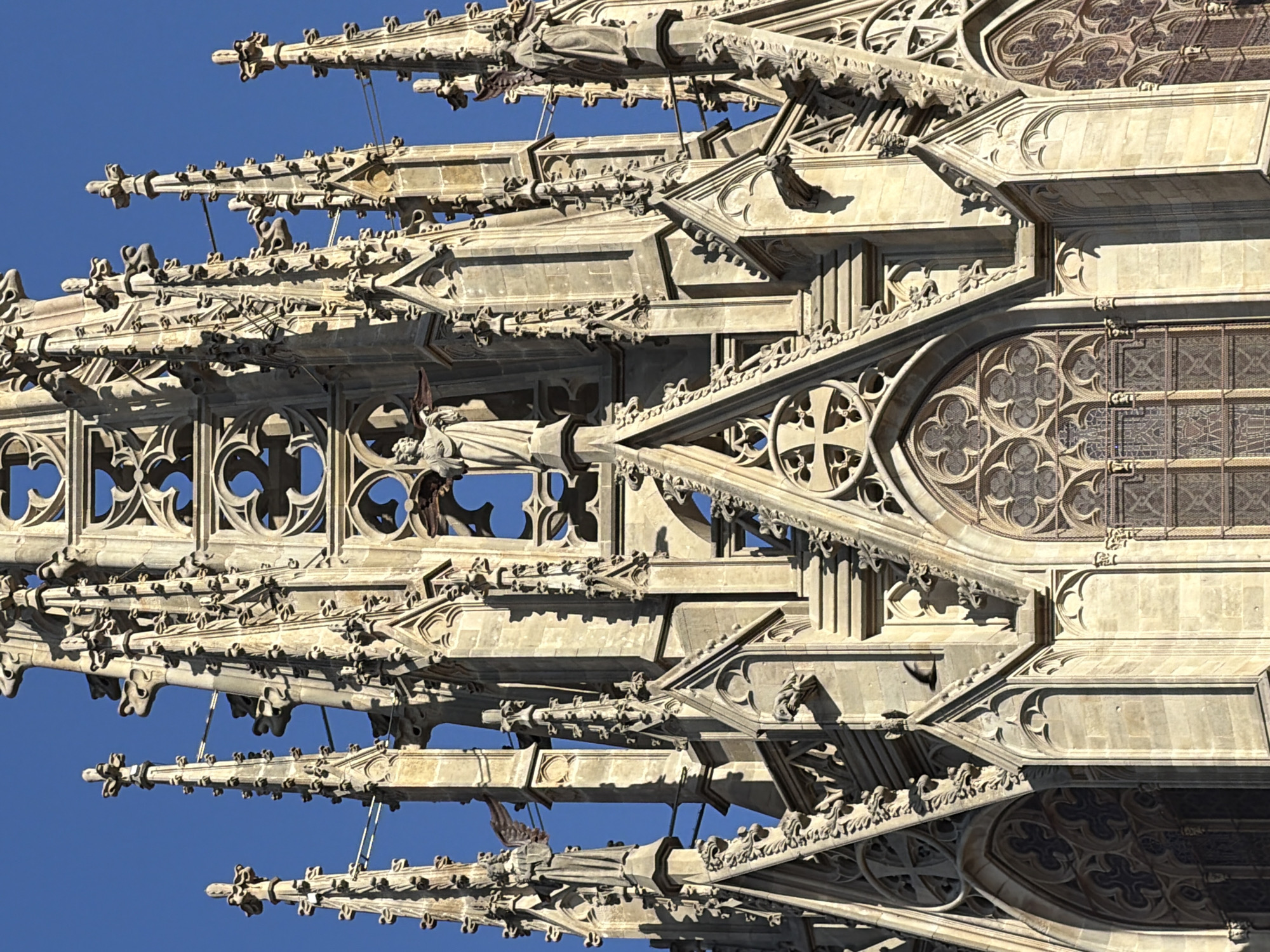 This media file captures a close-up, angled view of the intricate Gothic architecture of a cathedral against a clear, bright blue sky. The sunlight highlights the stonework, casting sharp shadows that emphasize the detailed carvings and decorative elements. The subjects are the architectural features themselves, including pointed spires, gargoyles, ornate tracery in arched windows, and decorative moldings. There are no people or active events depicted; the focus is solely on the static, detailed structure.
Notable details include the weathered appearance of the stone, suggesting age and exposure to the elements. The clear blue sky indicates a sunny day, likely midday or afternoon given the strong shadows. The location is identified as Barcelona, Spain, which is known for its stunning Gothic architecture, such as the Barcelona Cathedral. The image composition, with its overlapping layers of architectural detail and the upward tilt, creates a sense of grandeur and complexity. There is no visible text in the image.