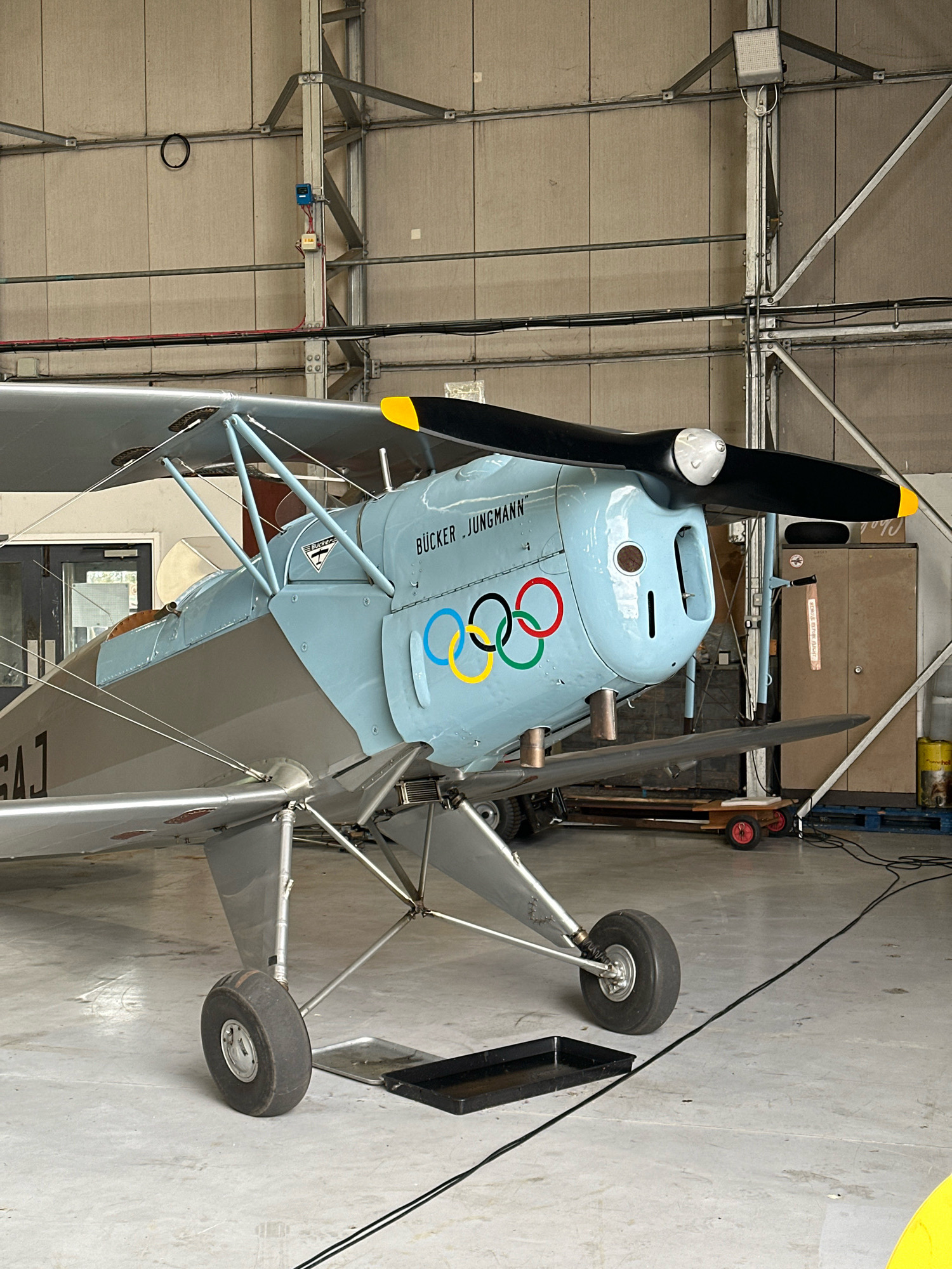 The image shows a light blue and silver biplane parked inside a hangar. The aircraft has the Olympic rings logo painted on its side and the name "BÜCKER JUNGMANN" written above it. The hangar appears to be a typical industrial building with concrete walls and a metal frame structure. The floor is concrete, and there are various objects scattered around, including wooden pallets, a yellow barrel, and some electrical equipment. The overall scene is well-lit, suggesting it's daytime. There are no people visible in the image.
The provided crops focus on different parts of the aircraft and the hangar interior, highlighting details such as the propeller, the cockpit area, the landing gear, and structural elements of the hangar.
Based on the provided image and the instruction to include the location in Cambridge, United Kingdom, it is not possible to definitively determine the specific location within Cambridge from the visual information alone. The image depicts a generic hangar setting, and there are no landmarks or identifying features that would pinpoint it to Cambridge.